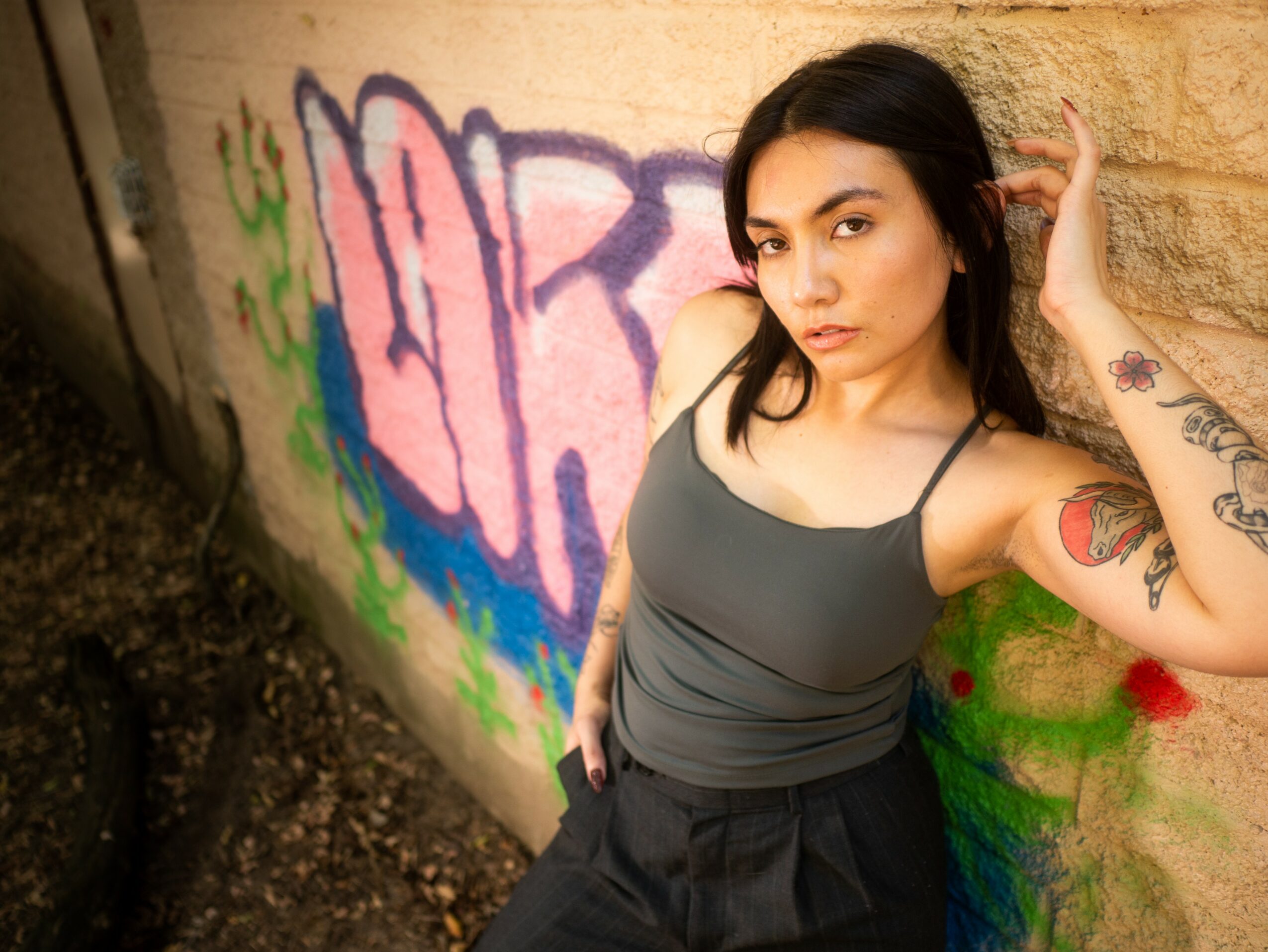 Young woman with dark hair and tattoos, wearing a gray tank top, leaning against a graffiti-covered wall.