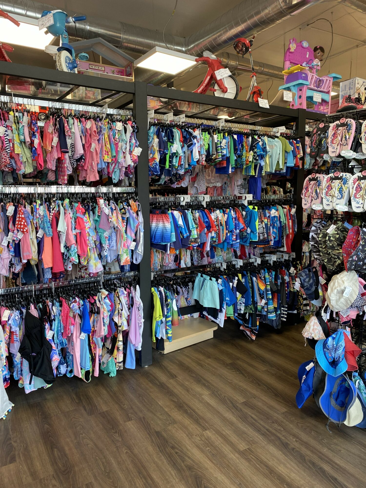 Children's clothing racks filled with colorful outfits in a store, with toys and hats displayed nearby, and a wooden floor.