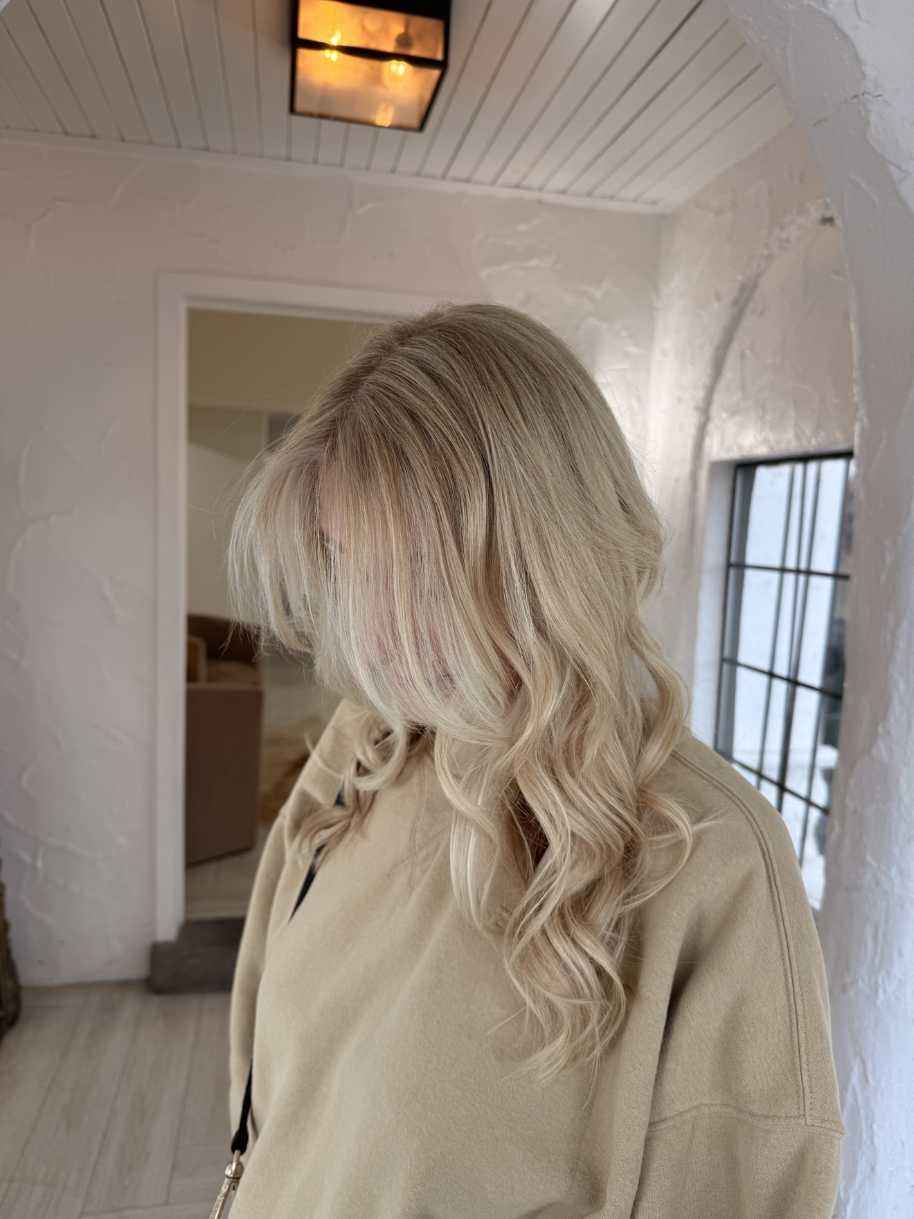 Person with long, wavy blonde hair wearing a beige jacket, facing away from the camera in a bright room.
