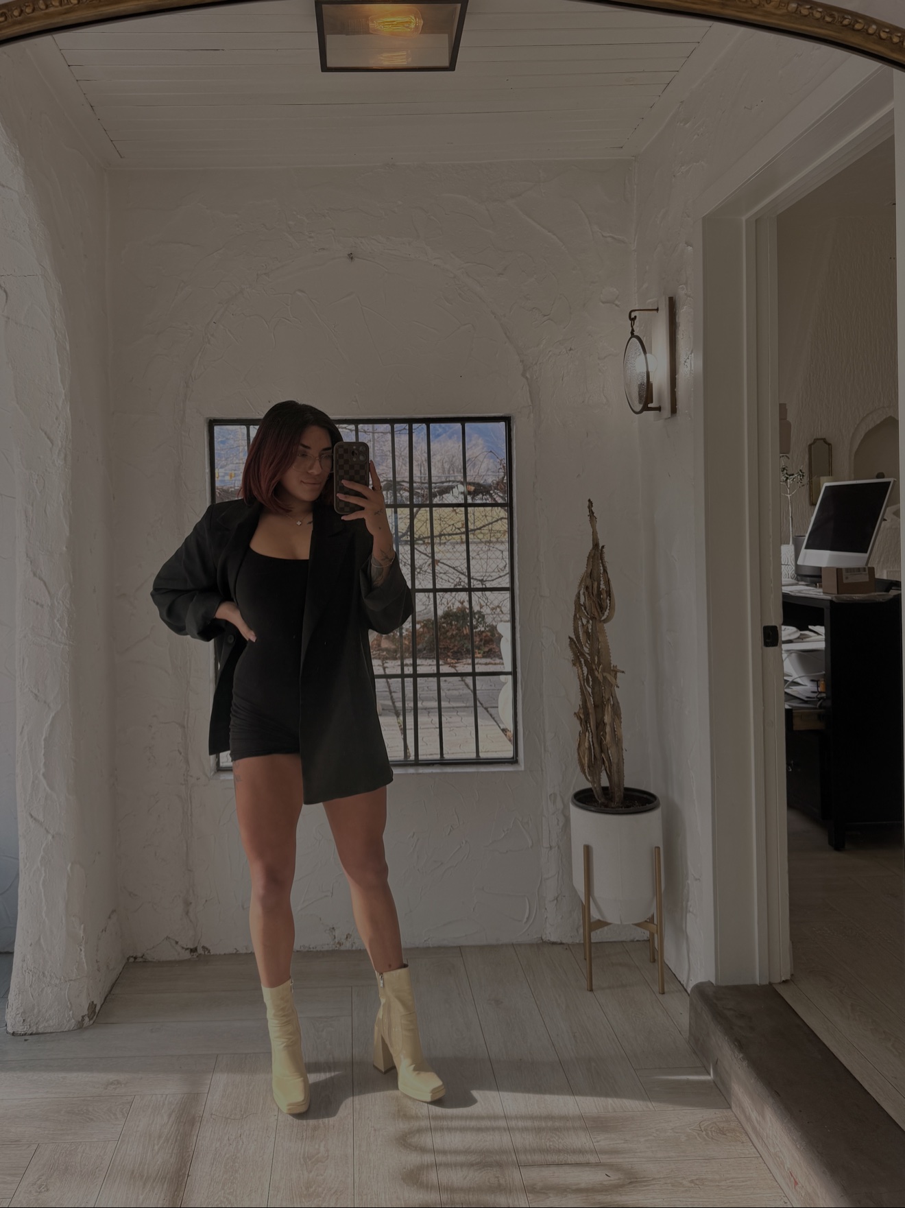 Person taking a mirror selfie in a room with a window, white walls, and a small table with a plant, wearing a black outfit and boots.