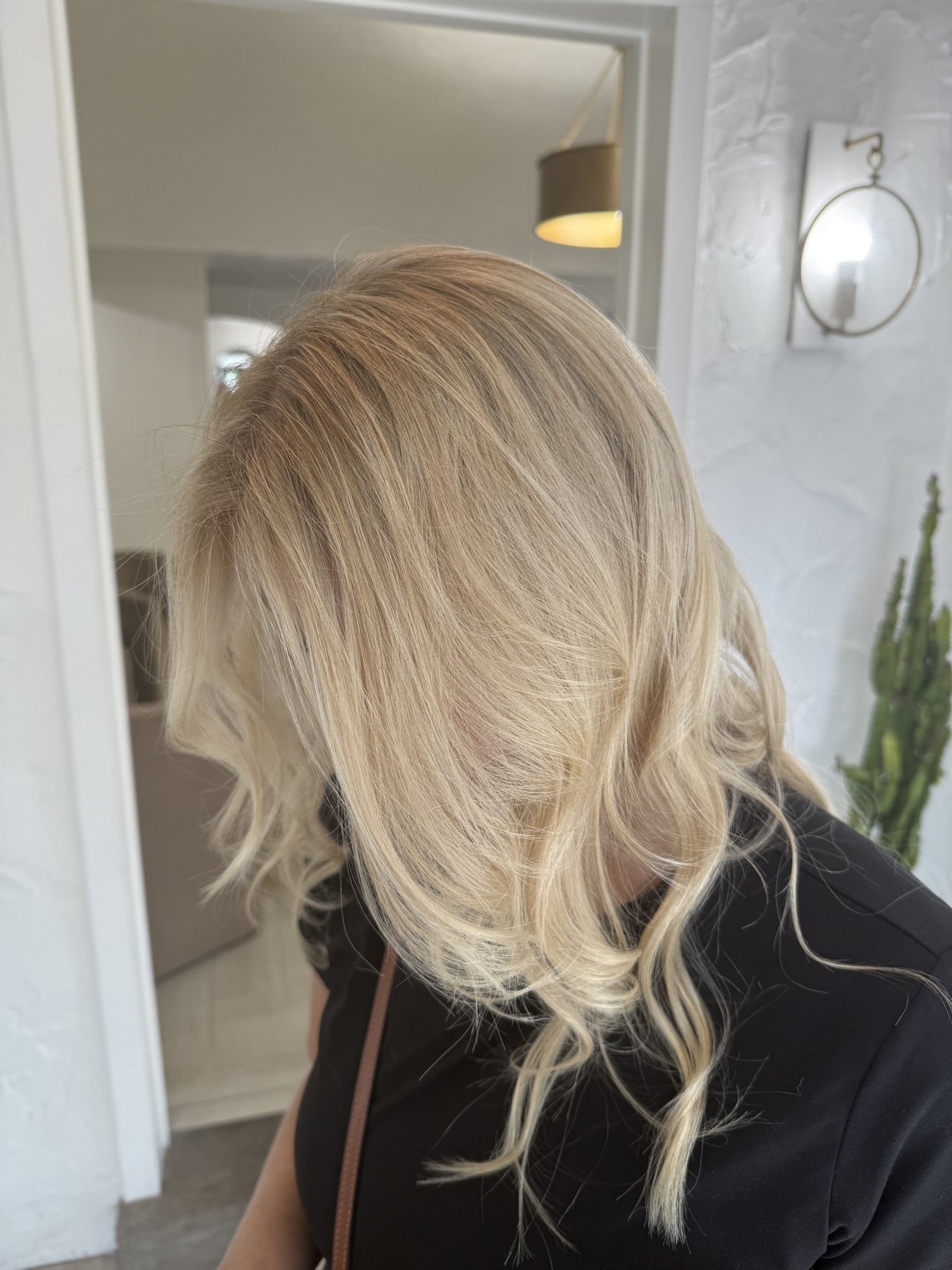 Blonde wavy hair partially covering face, indoors with white walls and a plant in the background.