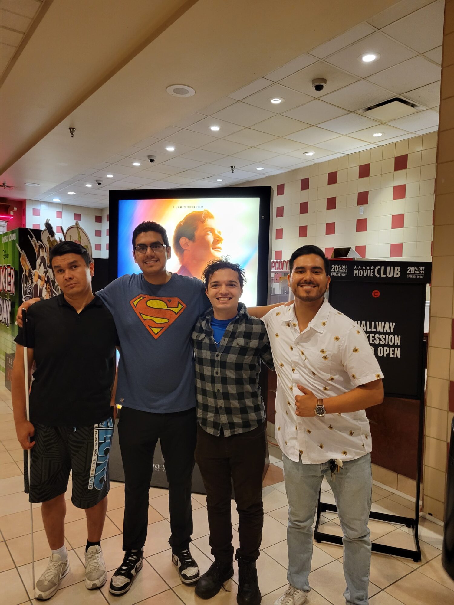 Four young men stand together in a shopping mall, smiling, with a large screen behind them displaying a person's face.