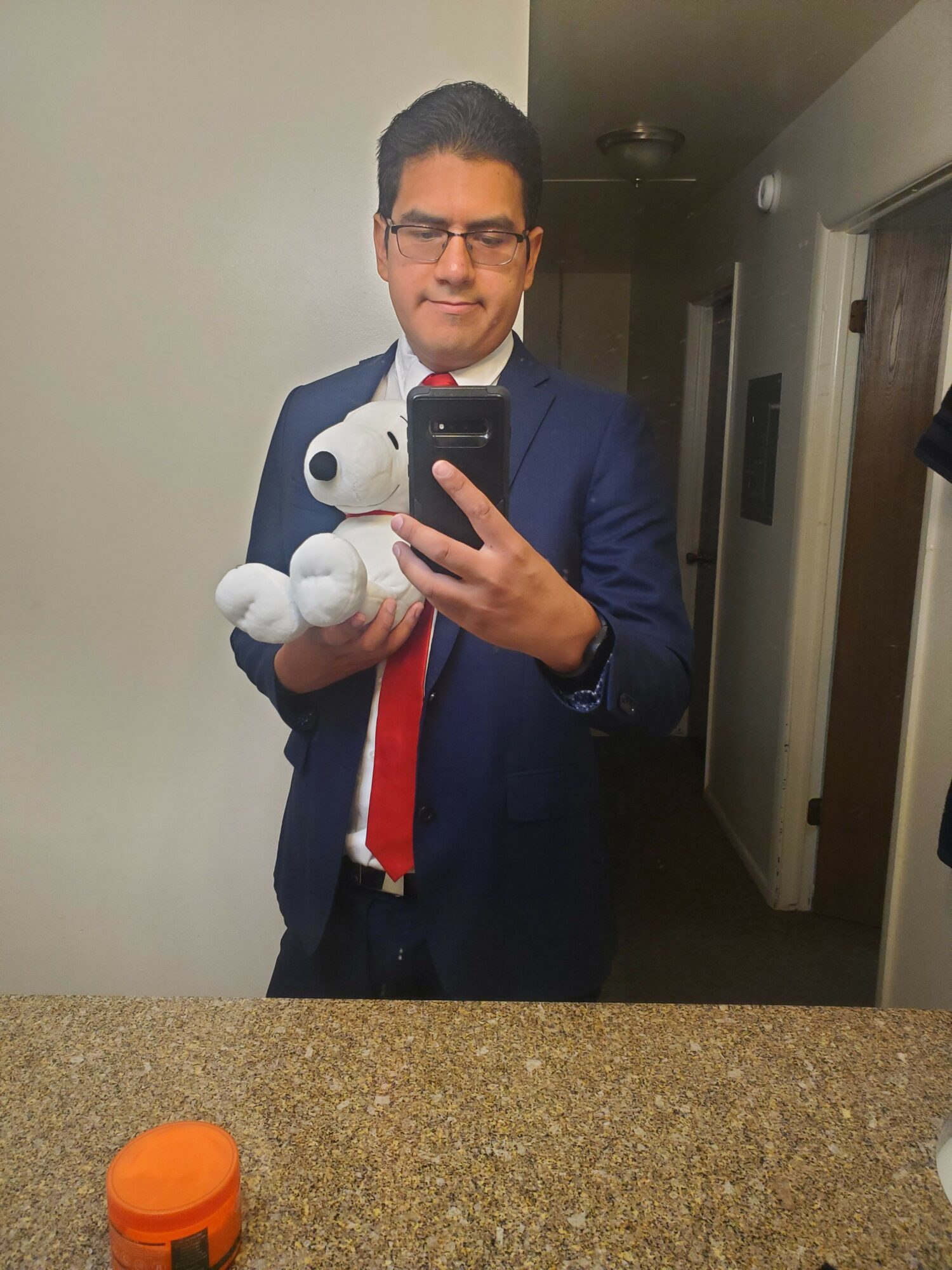 Man in suit taking mirror selfie, holding a stuffed animal, with a countertop and orange object visible.