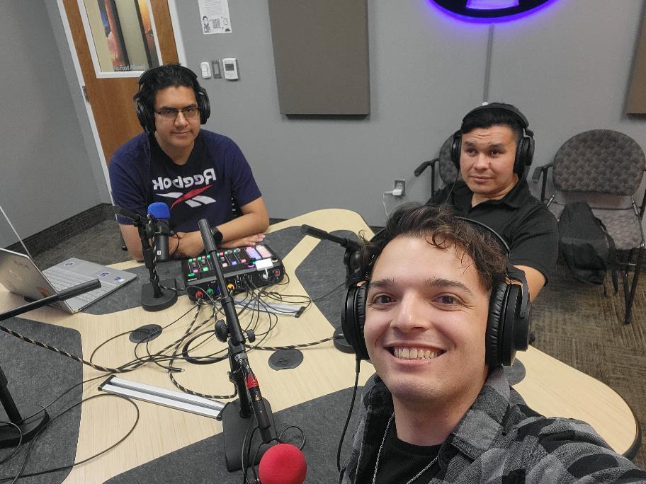 Three people in a recording studio with microphones and headphones, smiling at the camera.