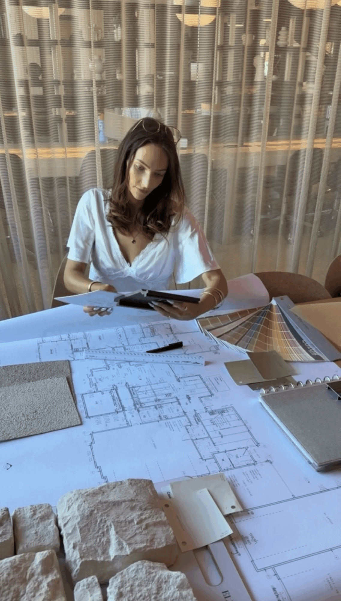 Woman sitting at a table with architectural plans, color swatches, and a laptop, holding a tablet, in front of a curtain.