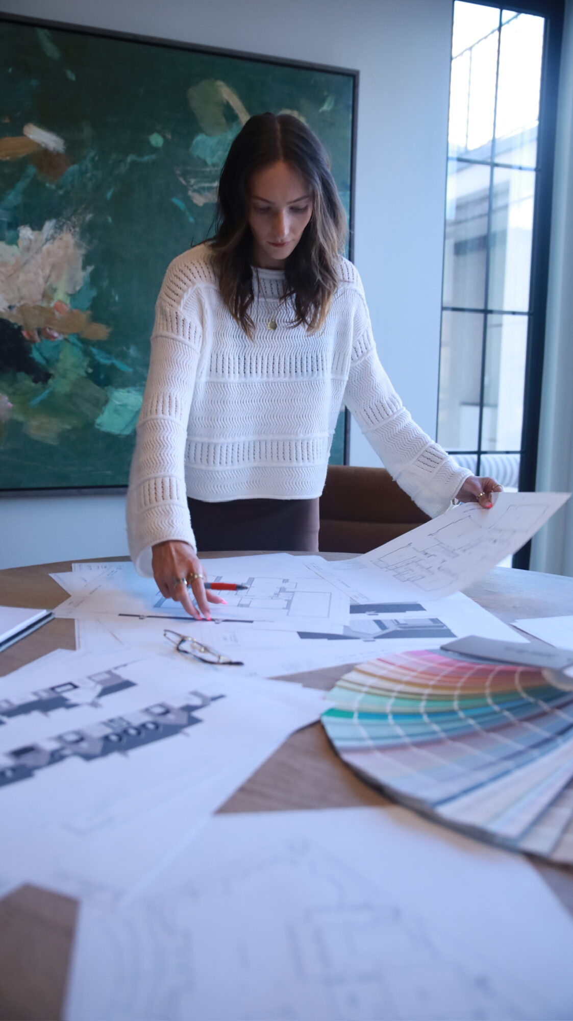 Woman in white sweater reviewing papers and color swatches on a table in a bright room with large windows.