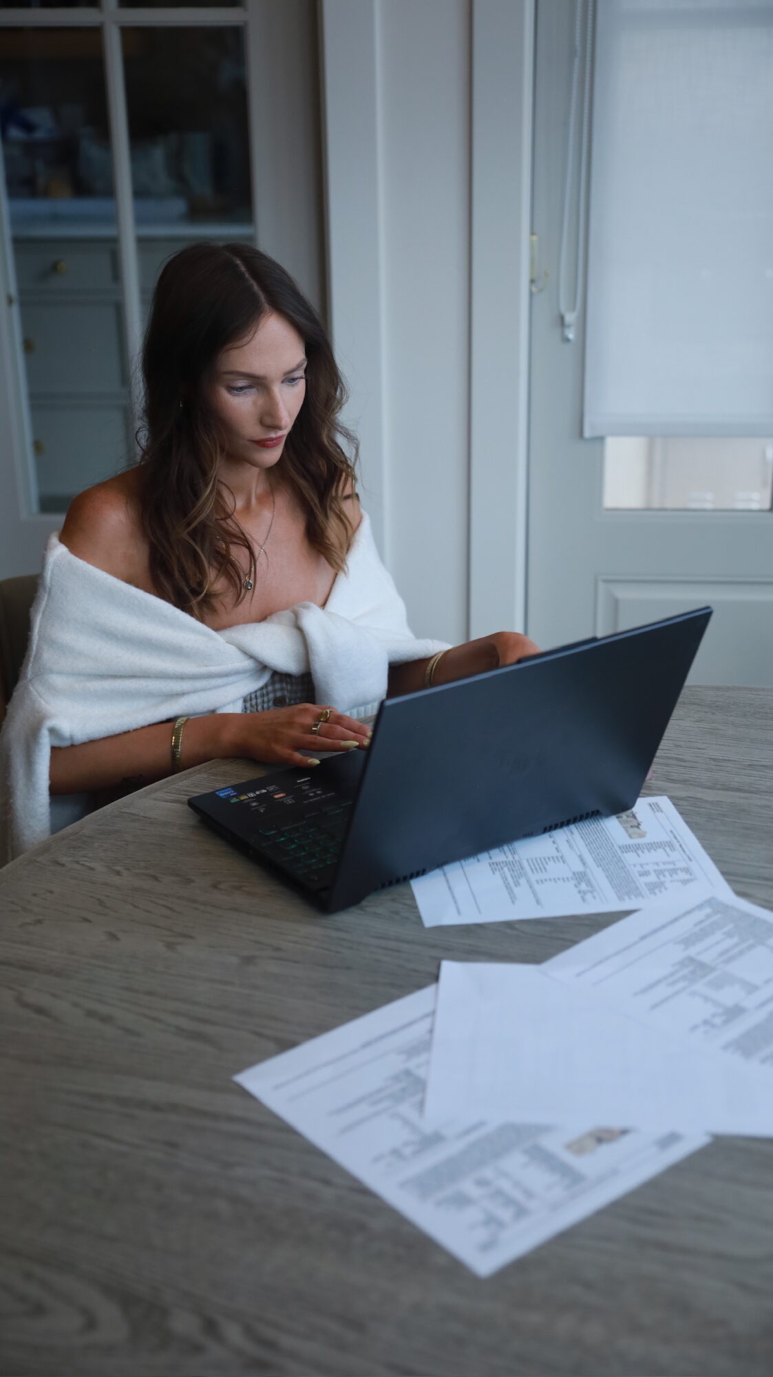 Woman with long hair working on a laptop at a table with papers, near a window with blinds.