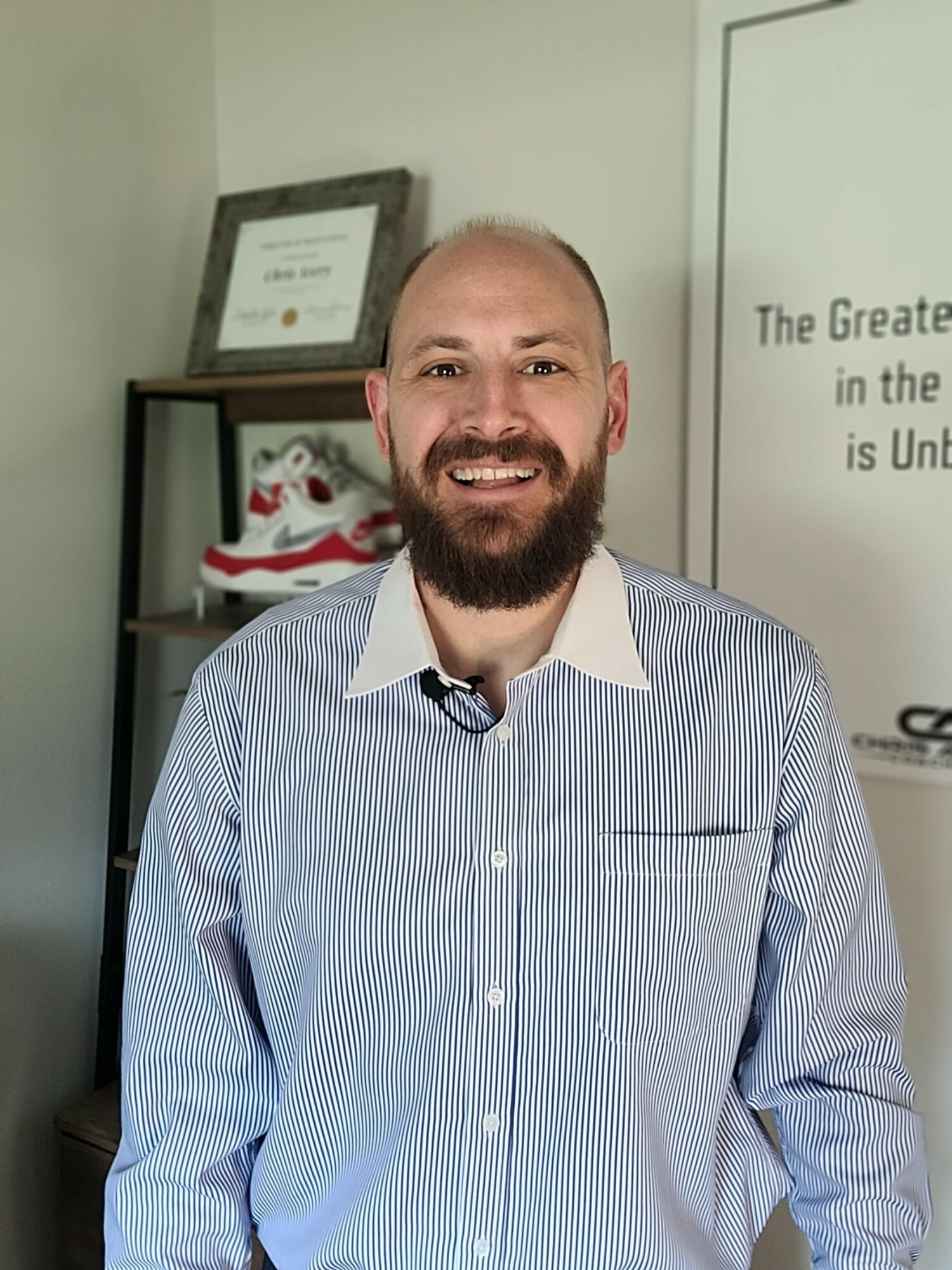 Smiling man with a beard and short hair, wearing a light blue striped shirt, standing indoors near a shelf and a framed certificate.