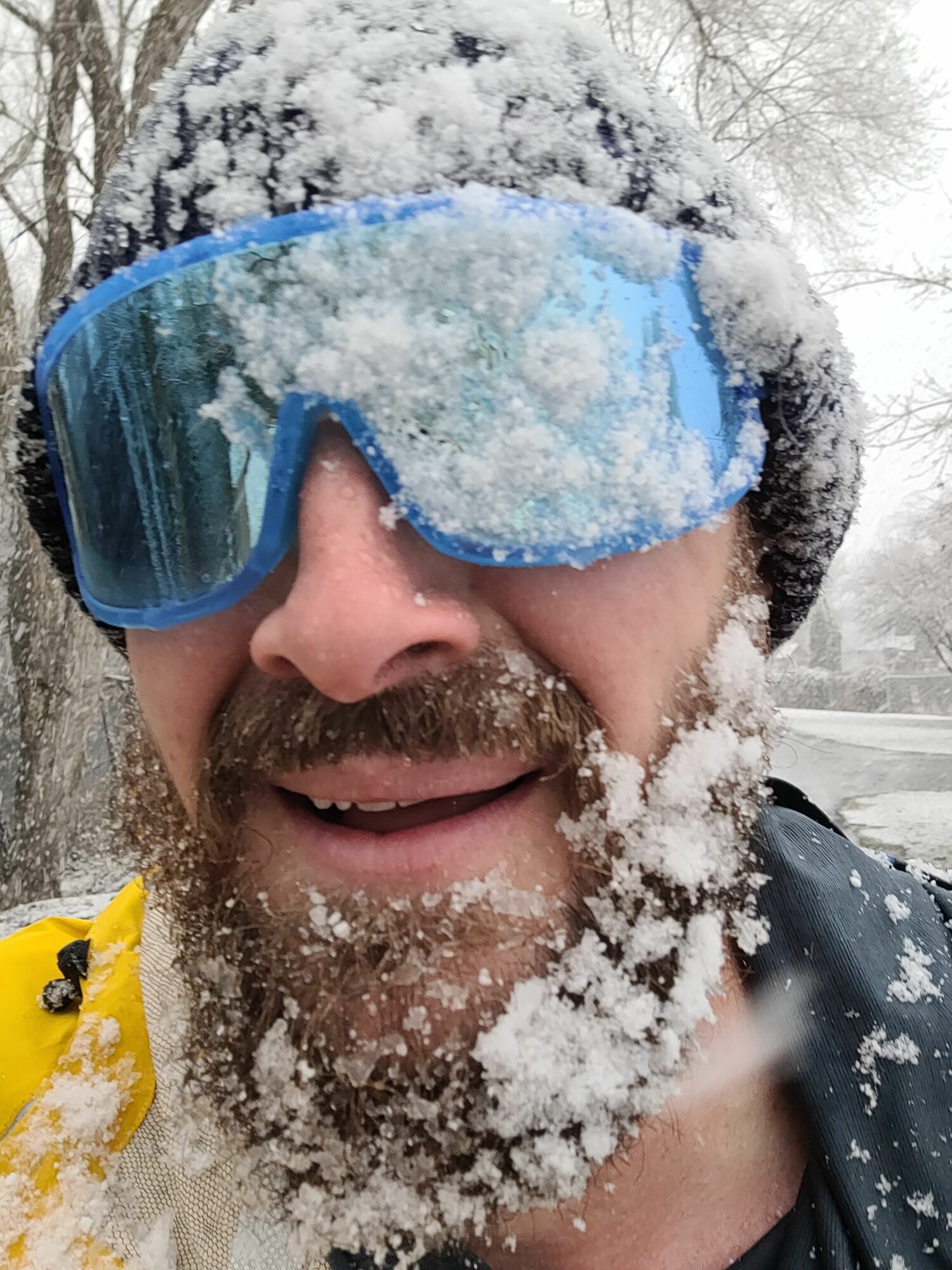 Person wearing blue snow goggles, smiling, surrounded by snow, outdoors in winter.