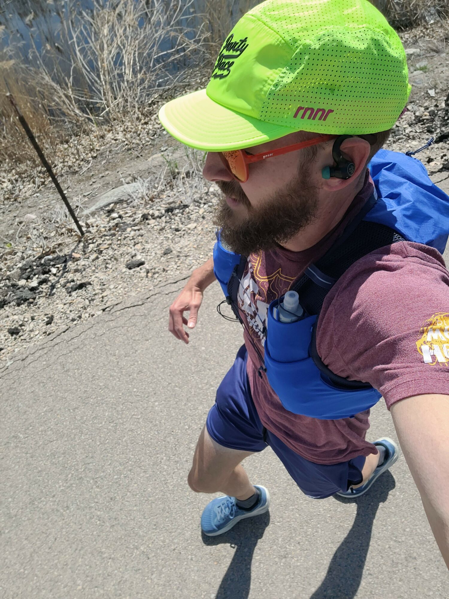 Person walking outdoors wearing a bright yellow cap, sunglasses, and casual clothing, with a backpack, on a sandy path.