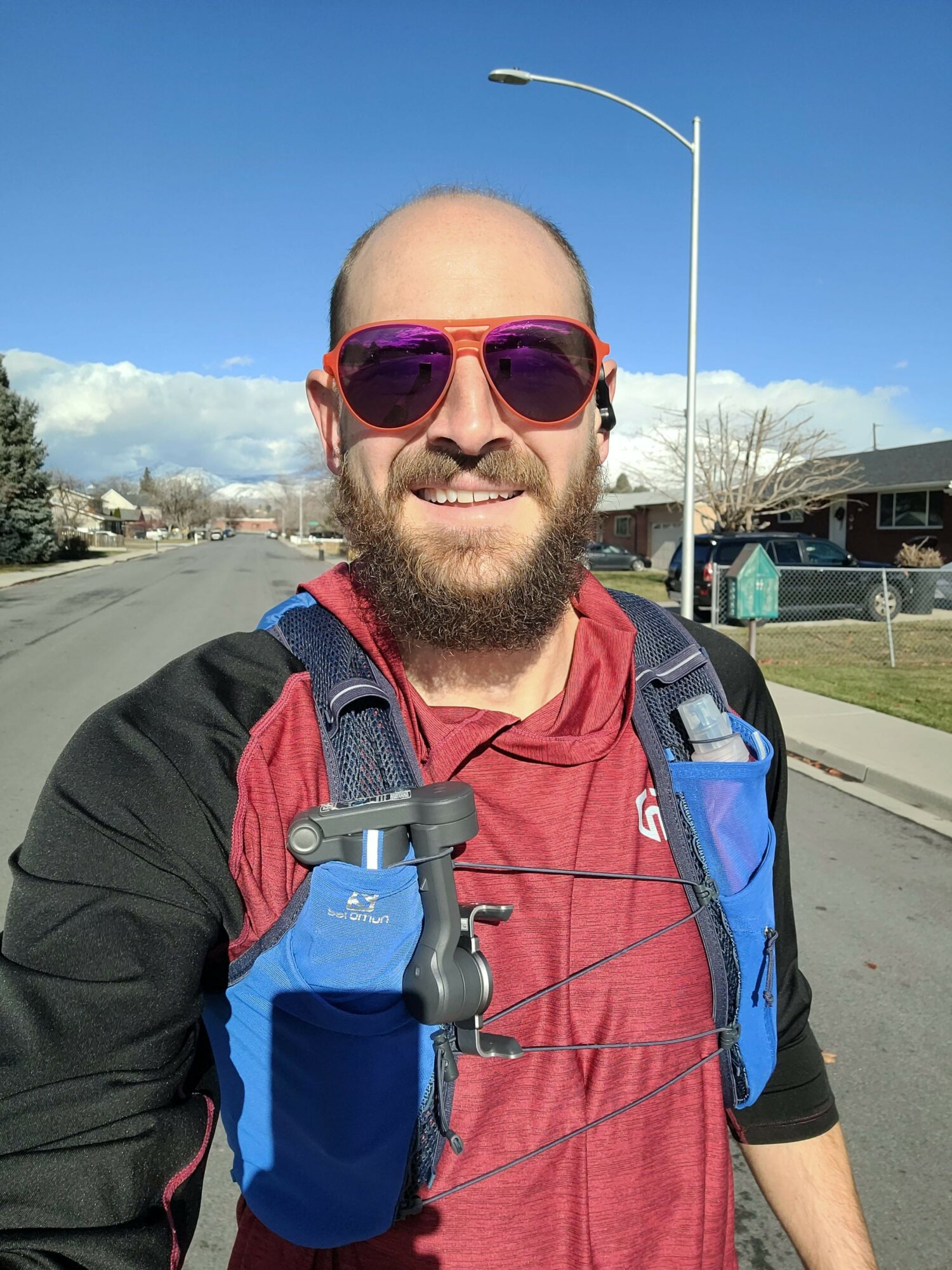 Man with sunglasses, beard, red shirt, and backpack outdoors on a sunny day.