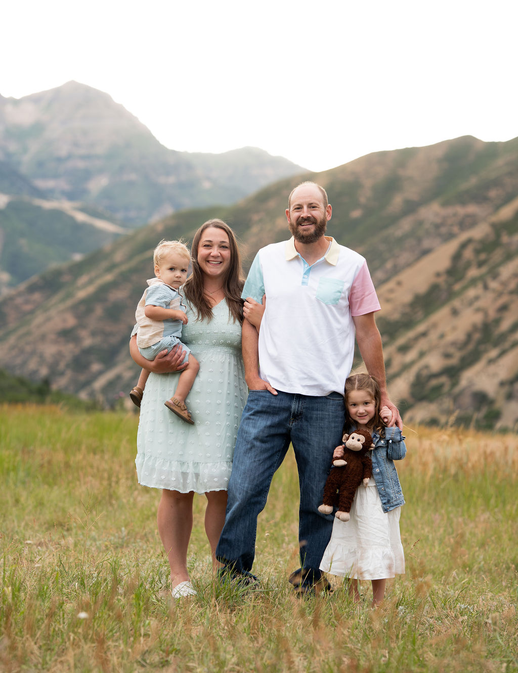 Family of four standing outdoors on grass with mountains in background, smiling, with two children, one held and one standing.