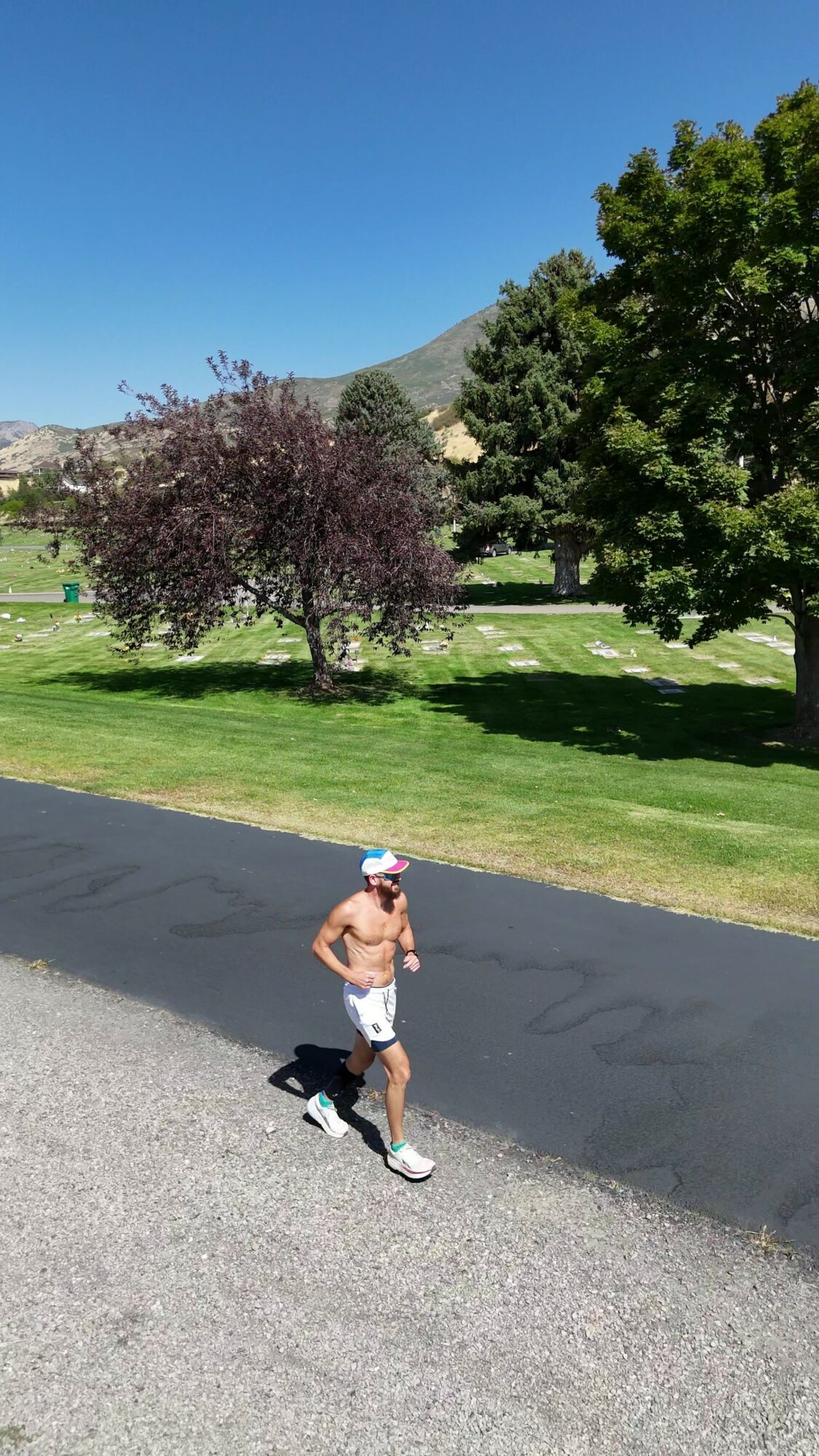 A person jogging on a paved path in a park with green grass, trees, and mountains in the background under a clear blue sky.