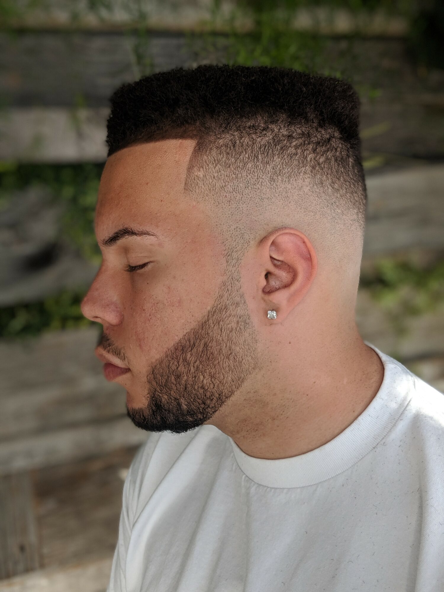Side profile of a young man with a high fade haircut, beard, and earring, outdoors with a wooden background.