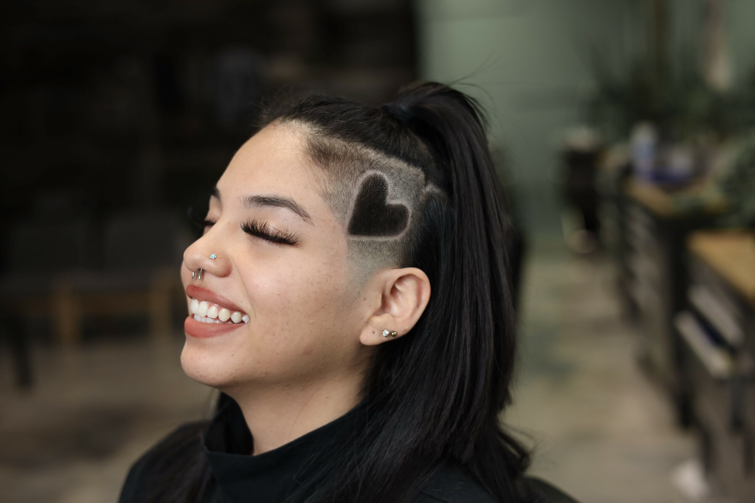 Smiling woman with a heart-shaped design shaved into her hair, in a casual indoor setting.