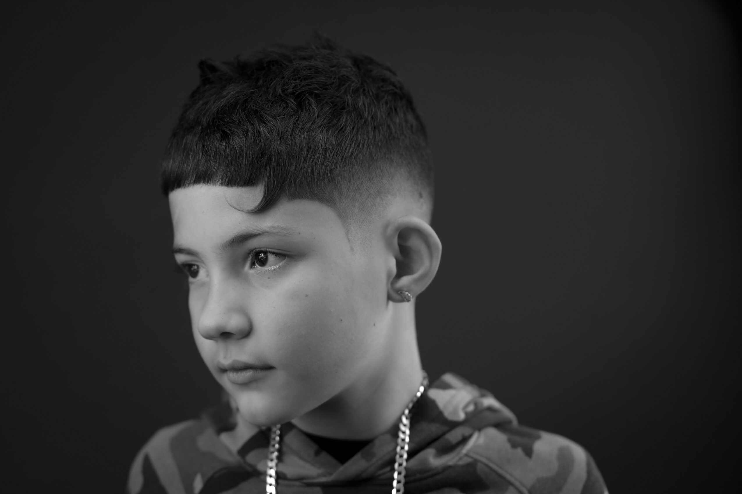 Portrait of a young boy with short hair, wearing a camouflage shirt and a chain necklace, looking to the side.