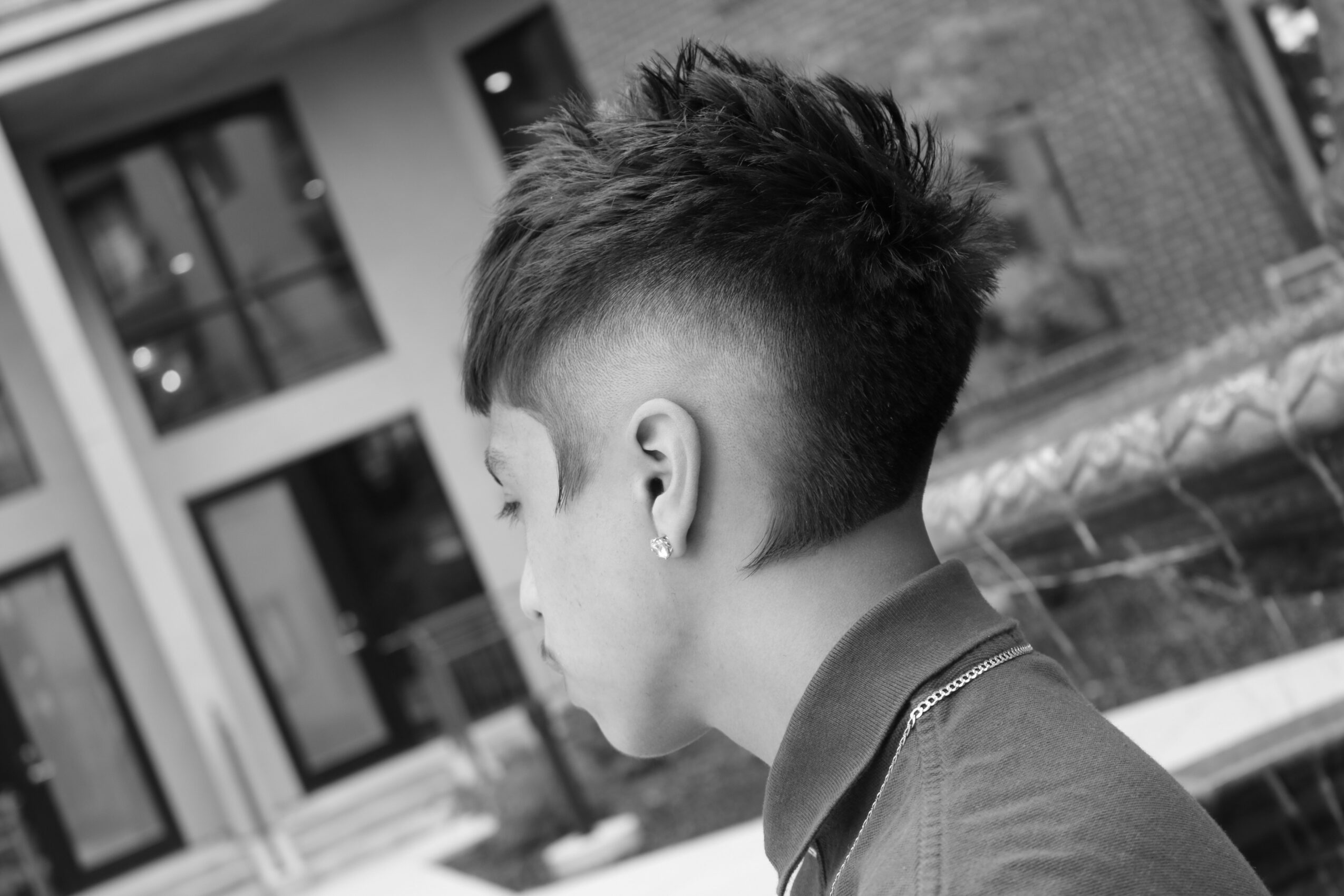 Side view of a young person with a short, spiked hairstyle, wearing a collared shirt, outdoors near a building.