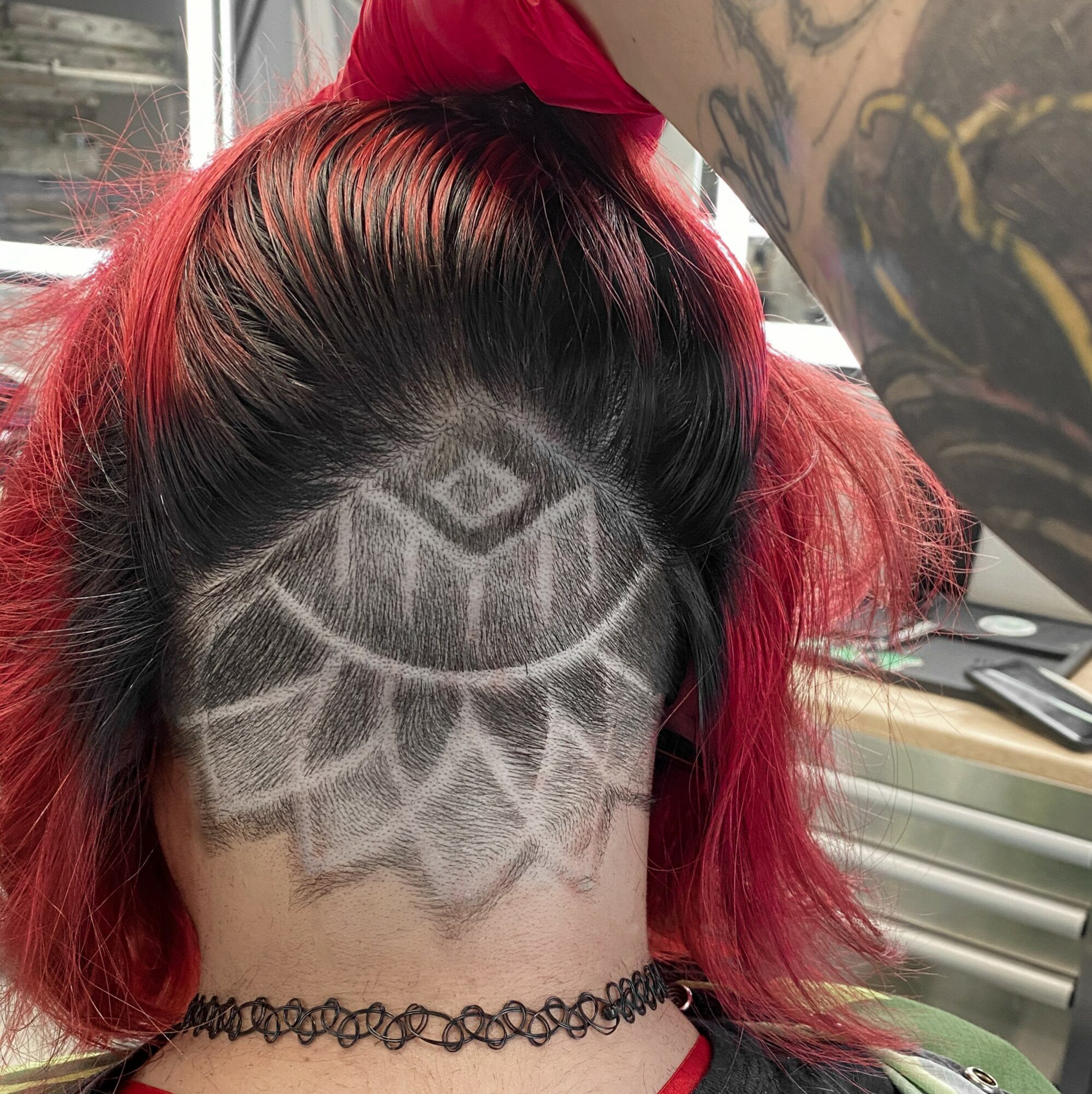Person with red and black hair showing a geometric shaved pattern on the scalp, wearing a black choker, in a room with a window and desk.