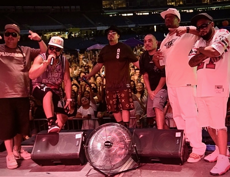 Group of six people on stage with a large crowd in the background, some holding microphones and wearing casual clothing.