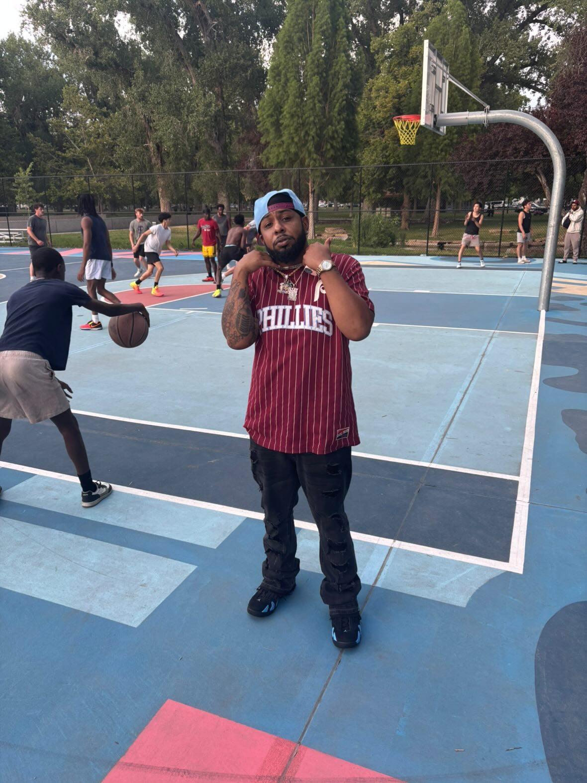 Person standing on a basketball court, holding a basketball, with others playing in the background, trees surrounding the court.