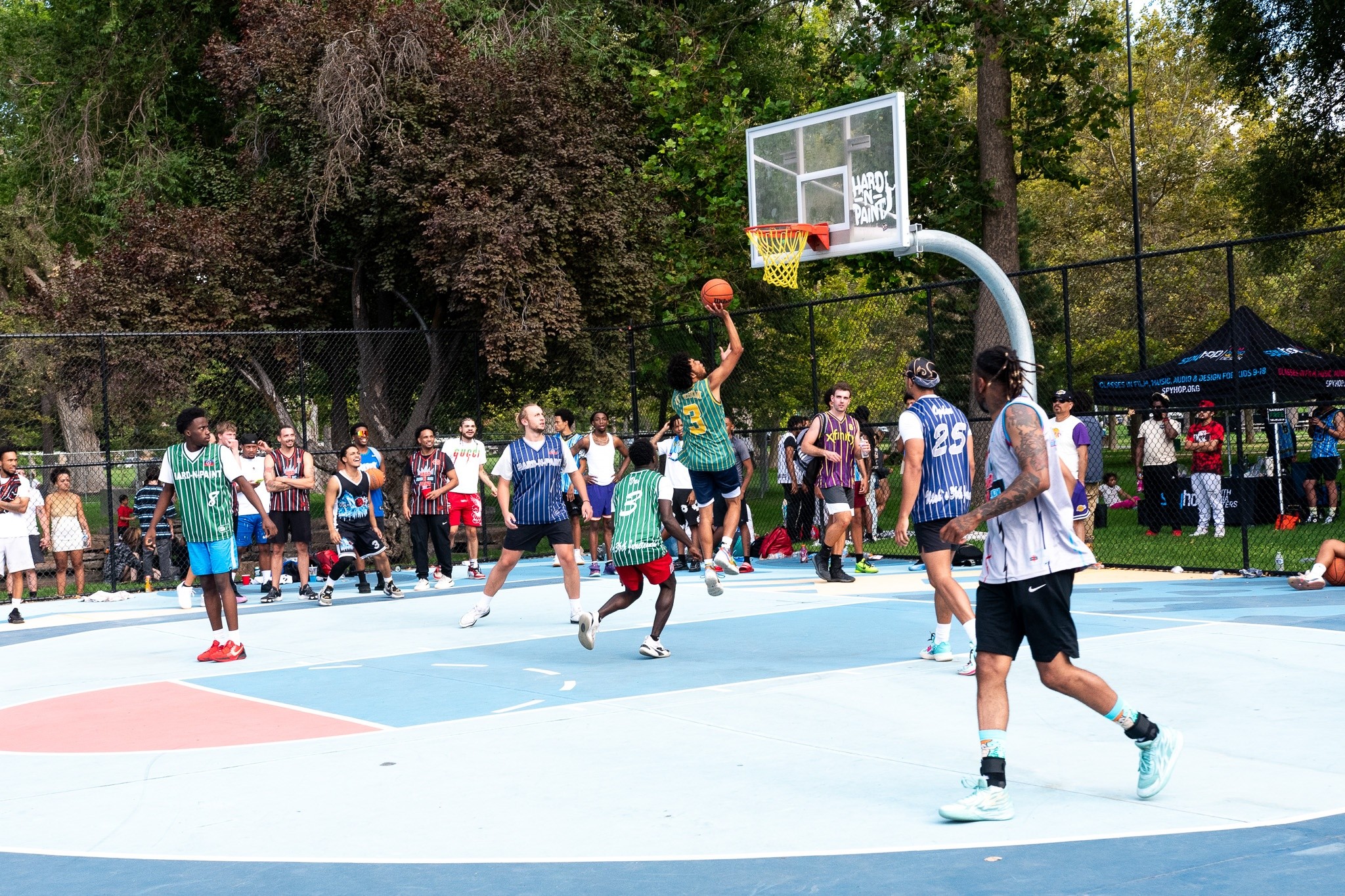 Group of people playing basketball outdoors on a court with trees in the background, some watching and others participating.