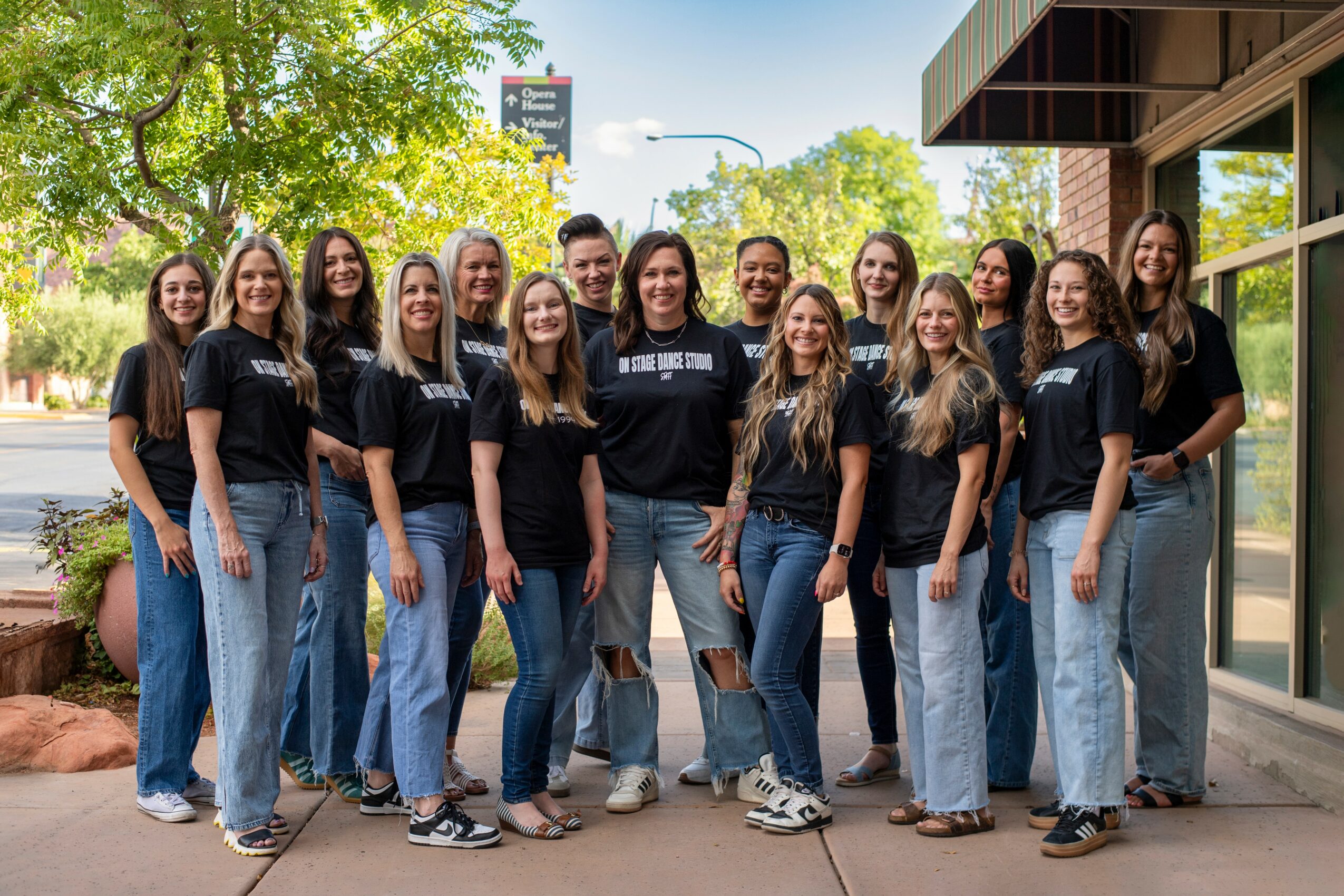 Group of fifteen women standing outdoors on a sidewalk, smiling, wearing black T-shirts and jeans, with trees and buildings in background.