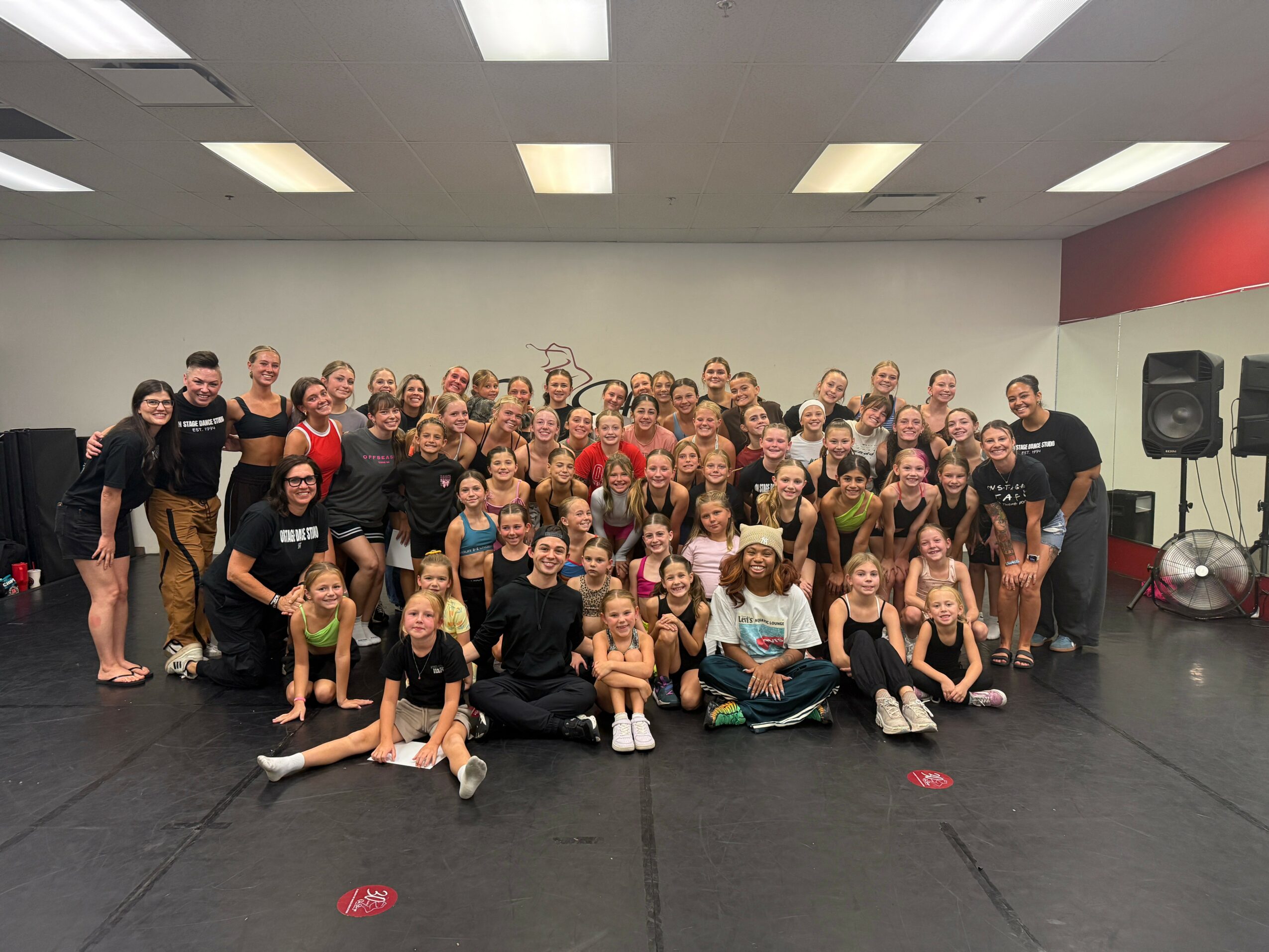 Group of children and adults in a dance studio, posing for a photo, some sitting and some standing, with a white wall and black floor.