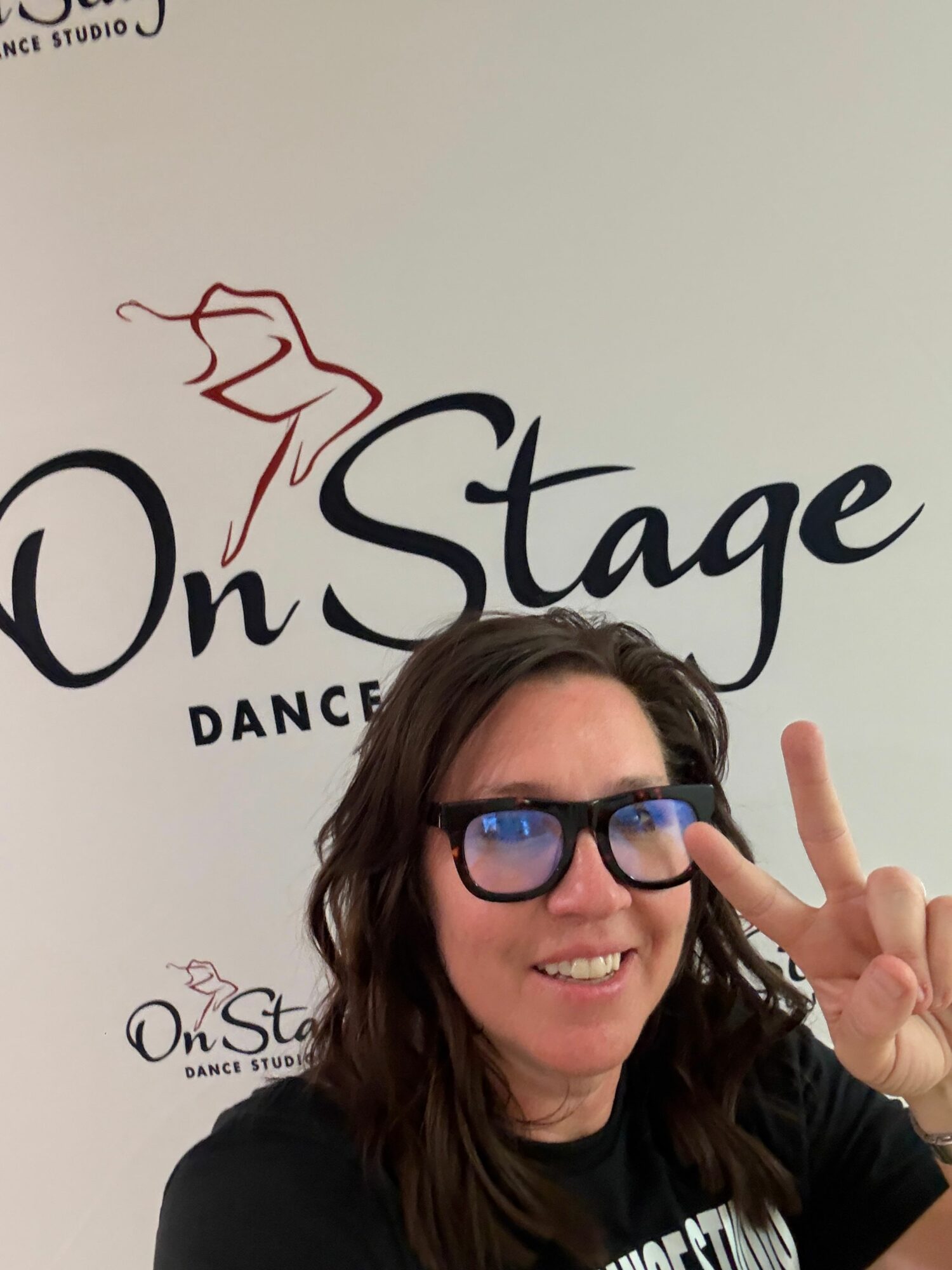 Woman with glasses smiling and making a peace sign near her face, standing in front of a dance studio sign.