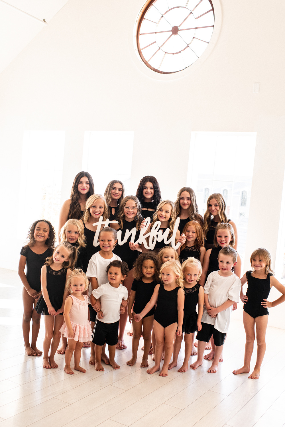 Group of children and adults posing indoors with a sign that says 'Thankful'.