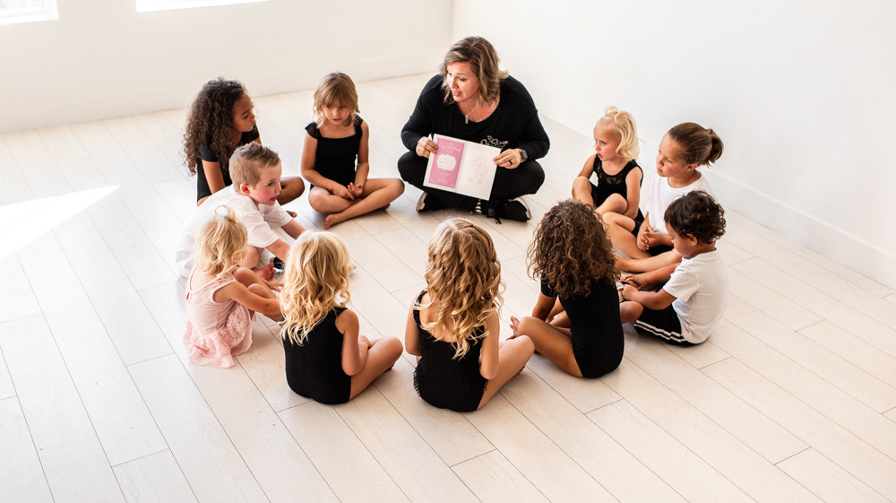 Adult woman reading a book to a circle of children sitting on a light-colored floor in a bright room.