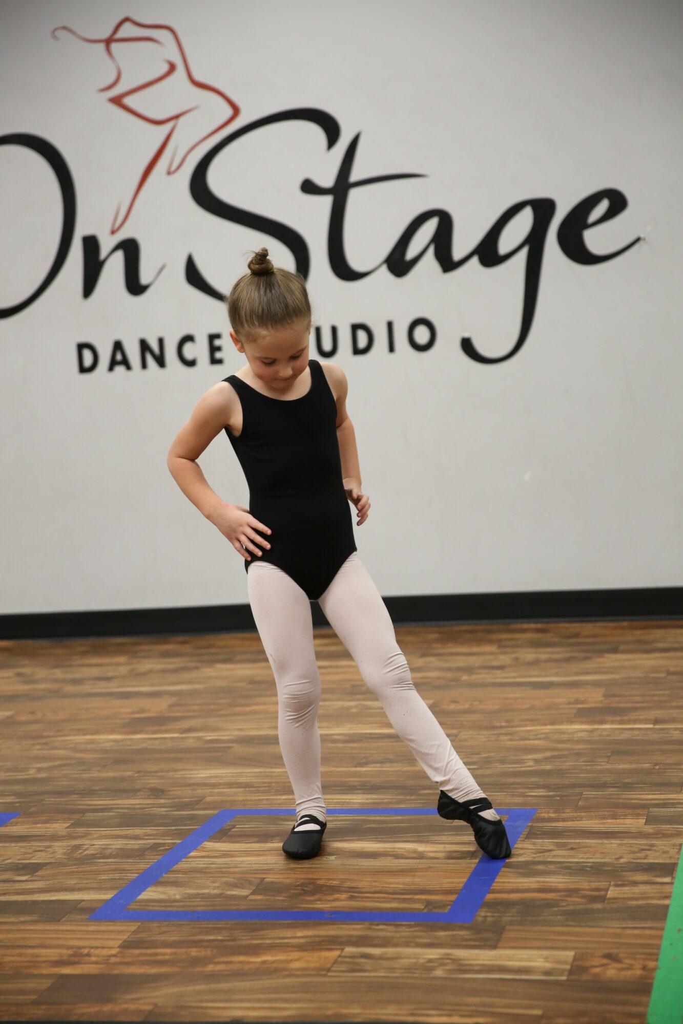 Young girl in dance attire standing inside a blue square on a wooden floor, with a dance studio sign in the background.
