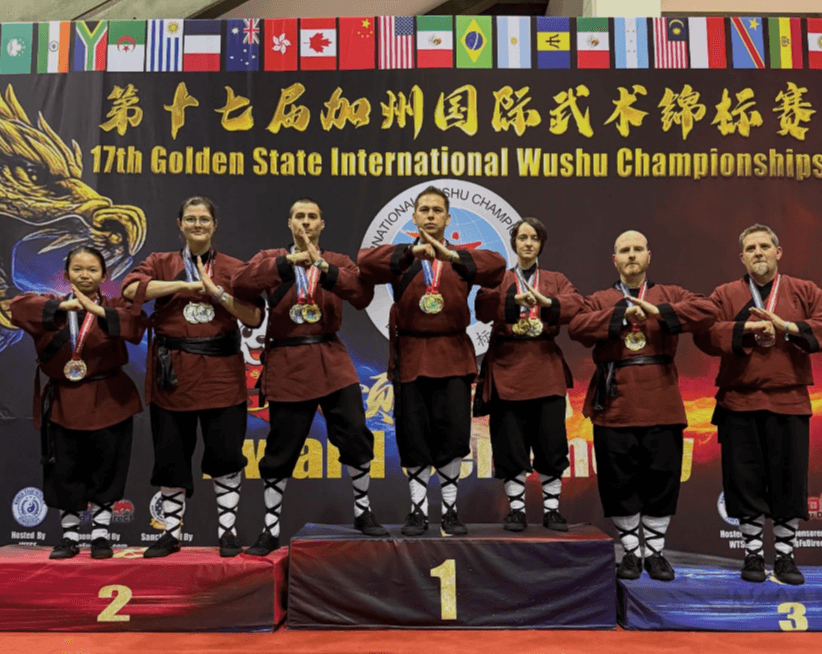 Seven martial artists stand on a podium with medals, holding hands, in front of a banner for the 17th Golden State International Wushu Championships.