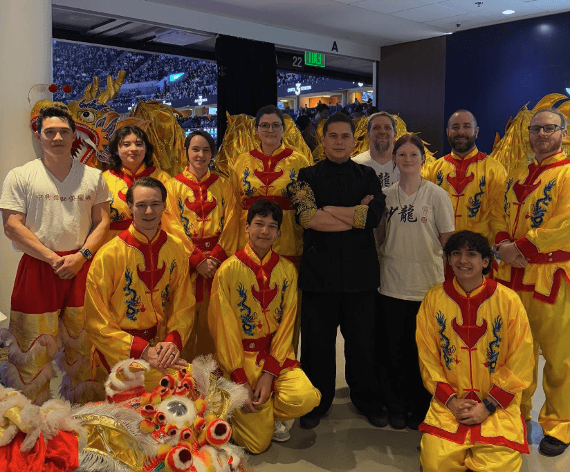 Group of people in traditional Chinese lion dance costumes and casual clothing indoors, with a window showing a cityscape at night.
