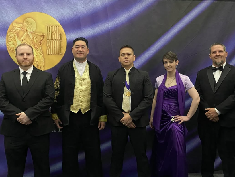Five people in formal attire standing in front of a dark backdrop with a gold emblem, posing for a photo.