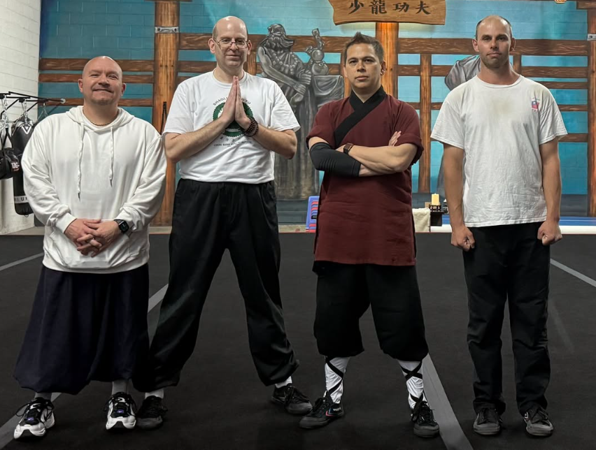 Four men standing in martial arts uniforms and casual clothing in a dojo, with a blue wall and martial arts decorations behind them.