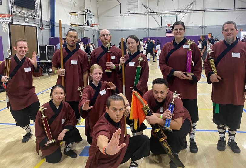 Group of ten people in maroon and black costumes holding swords and props in a gymnasium, smiling and posing.
