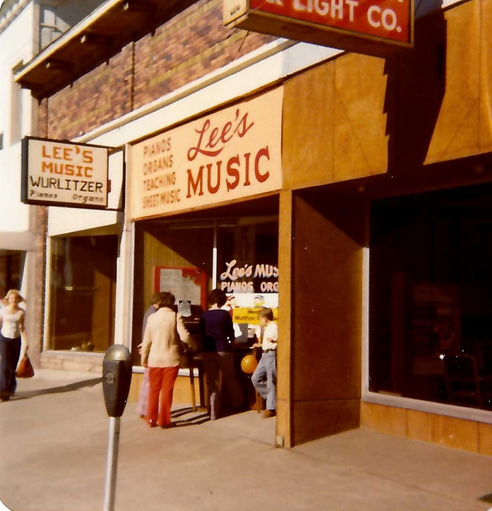 Group of people standing outside a music store with signs reading 'Lee's Music' and 'Pianos, Organs, Sound Equipment'.