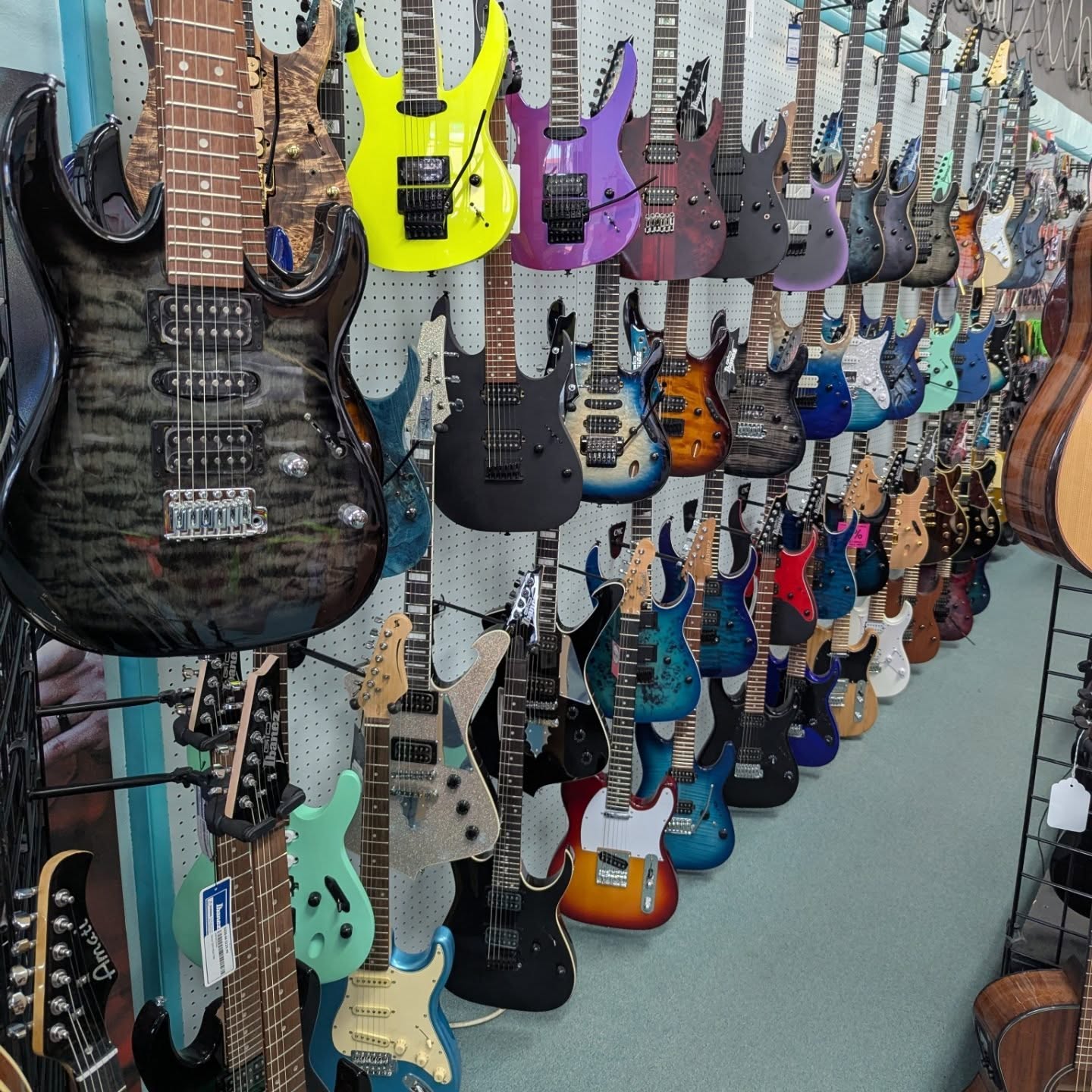 Display of various electric guitars in different colors and styles hanging on a wall, with some on stands and a few on the floor.