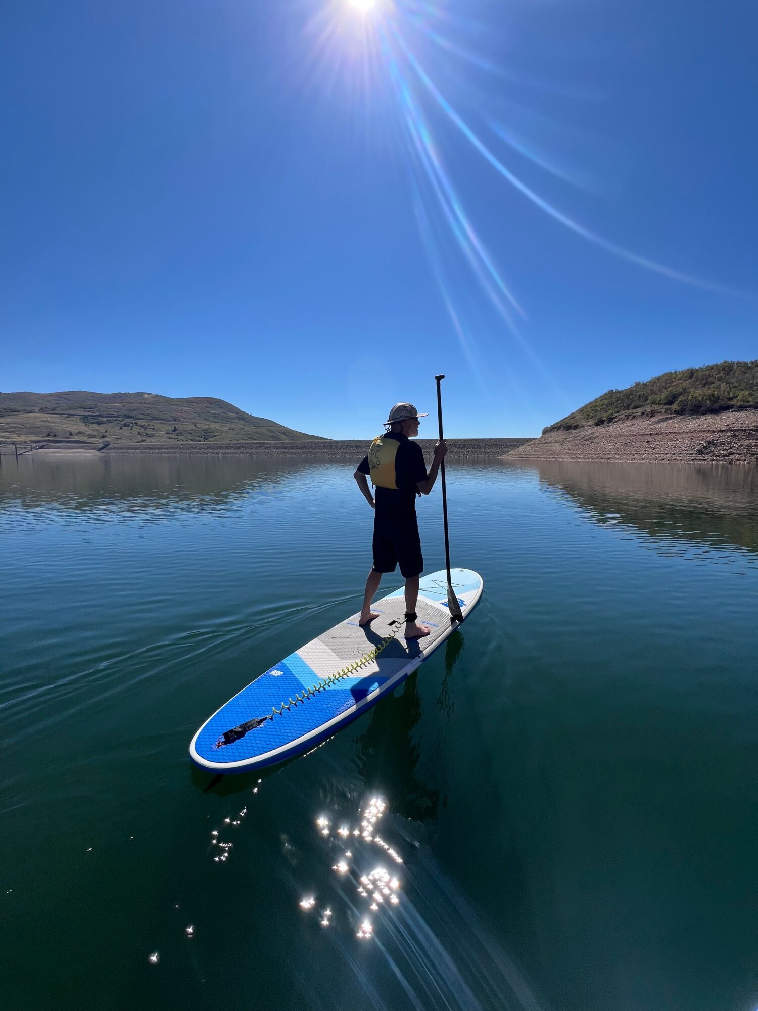 Person standing on a paddleboard on calm water near a rocky shoreline under a bright sun.
