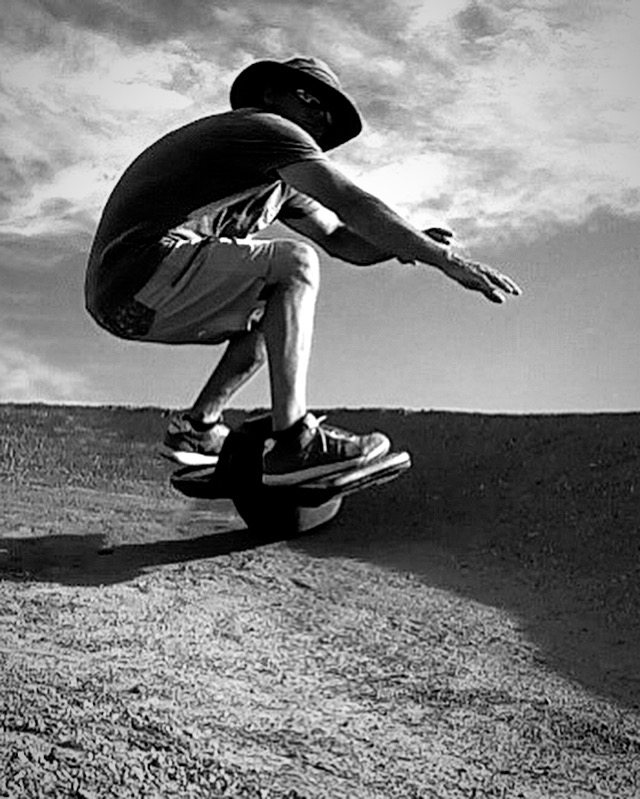 A person wearing a hat crouches in midair above a paved surface, with one arm extended.