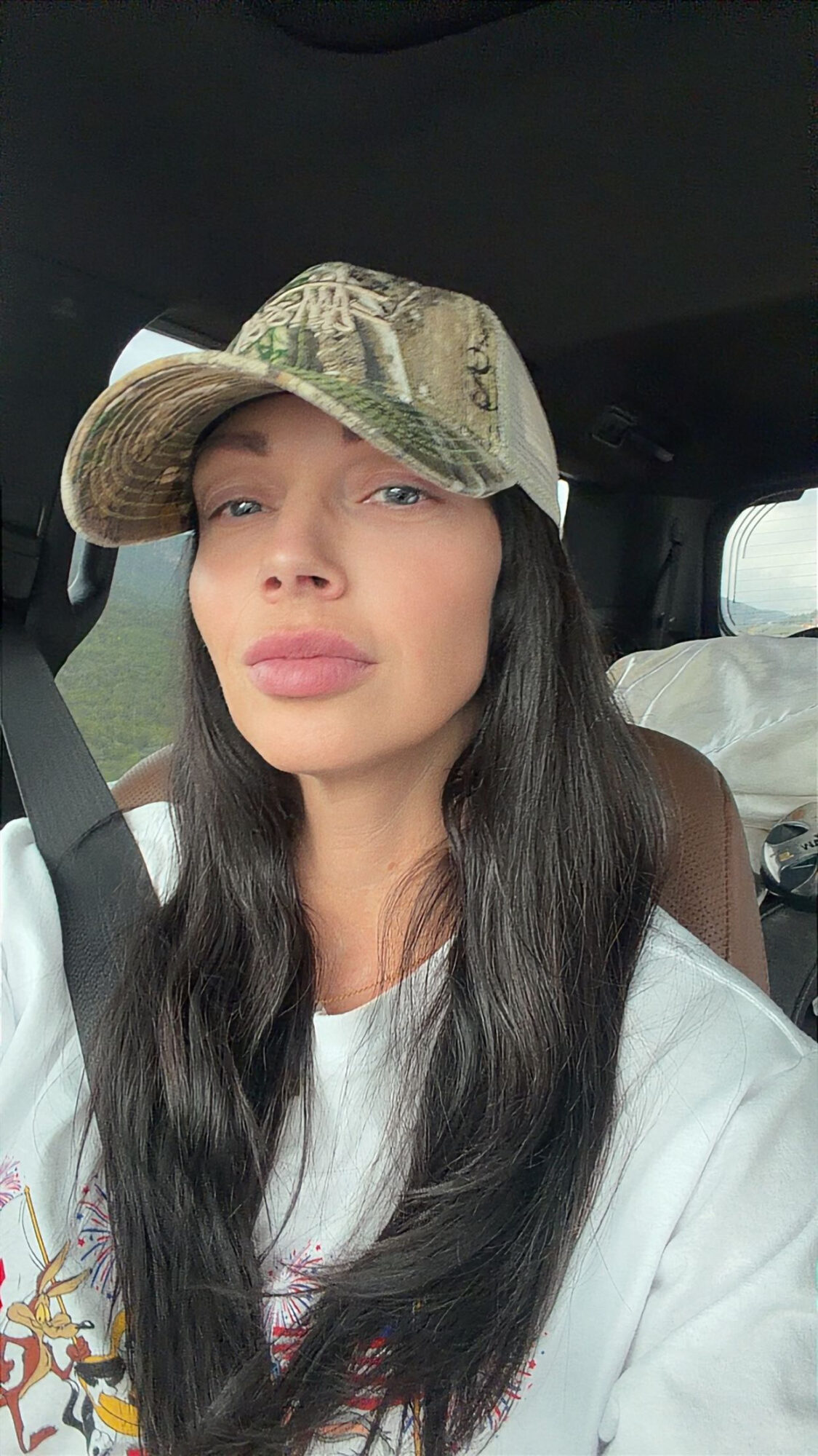 Person with long dark hair wearing a camouflage cap and white shirt, seated in a car.