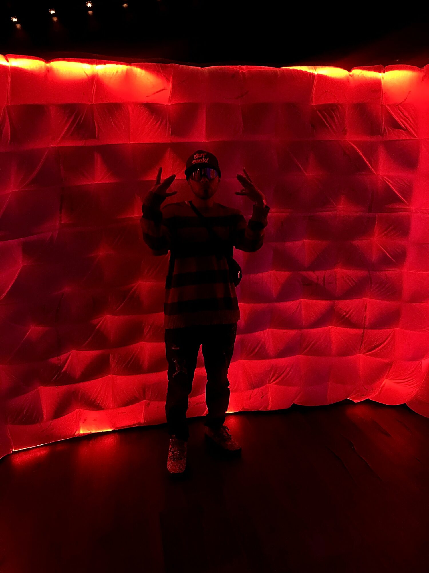Person standing in front of a red, textured wall, making peace signs with both hands, silhouette visible against the illuminated background.
