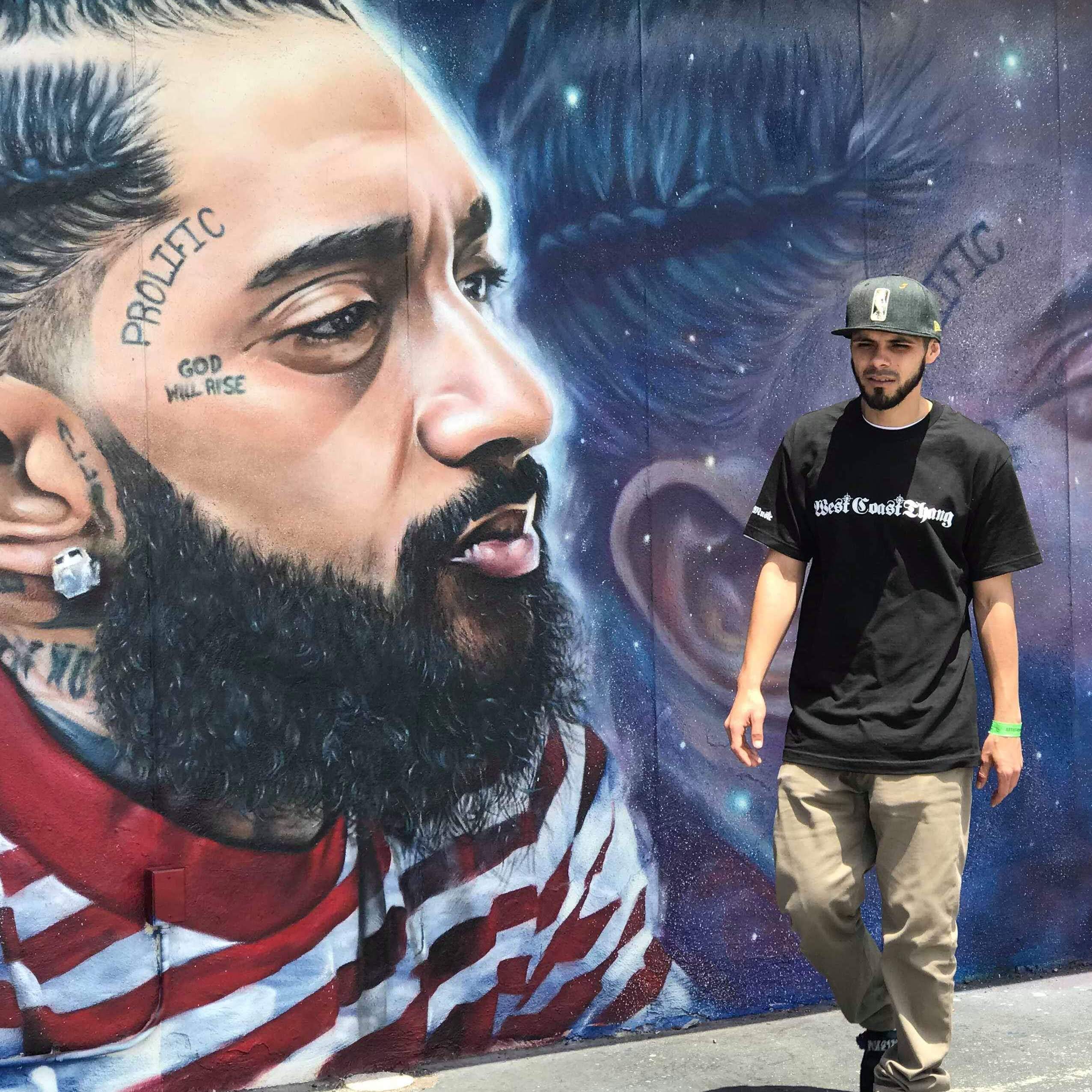 Person standing in front of a large mural of a bearded man with tattoos on his face, wearing a black t-shirt and cap.