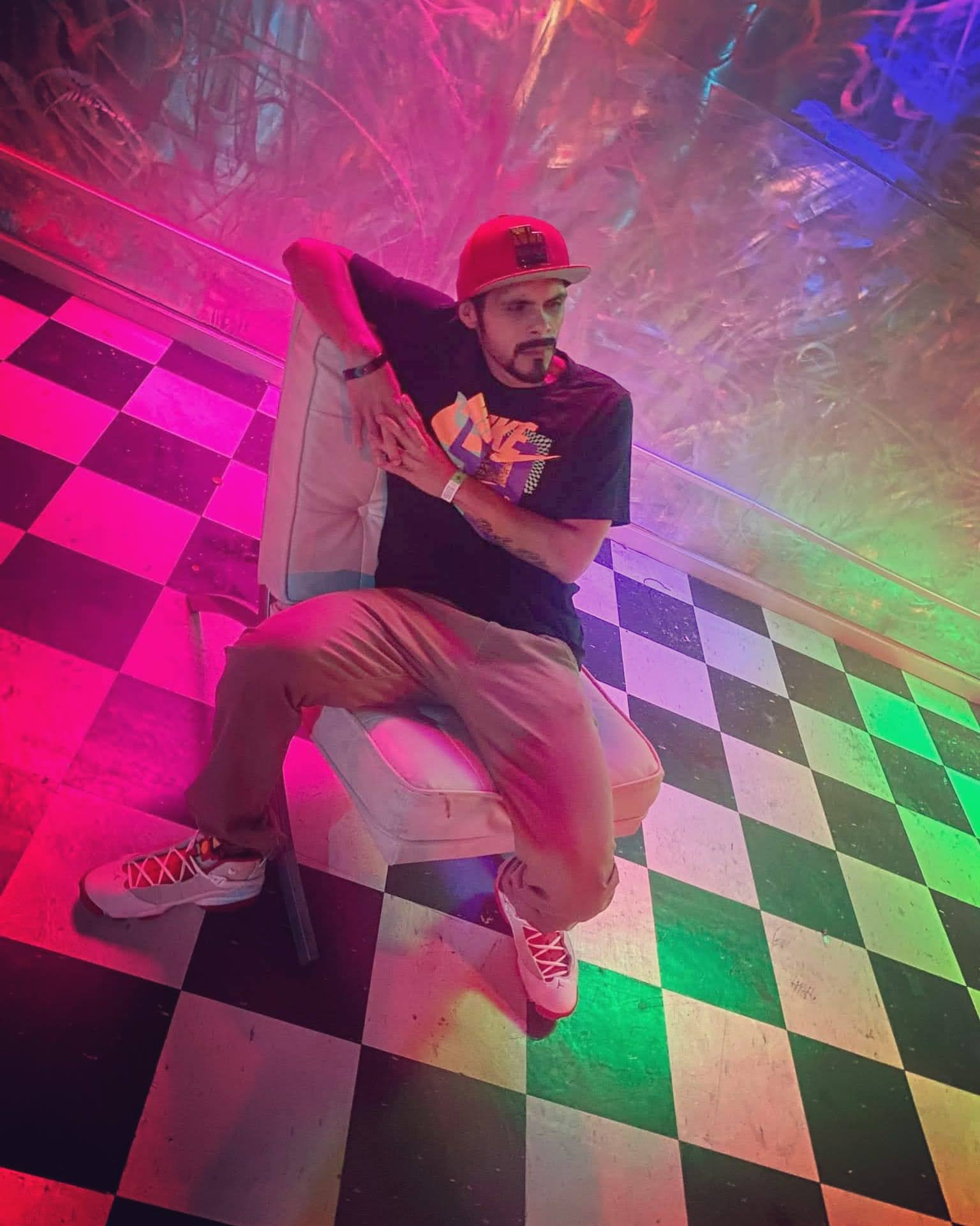 Person sitting on a chair with colorful lighting and checkered floor, wearing a cap, t-shirt, and sneakers.