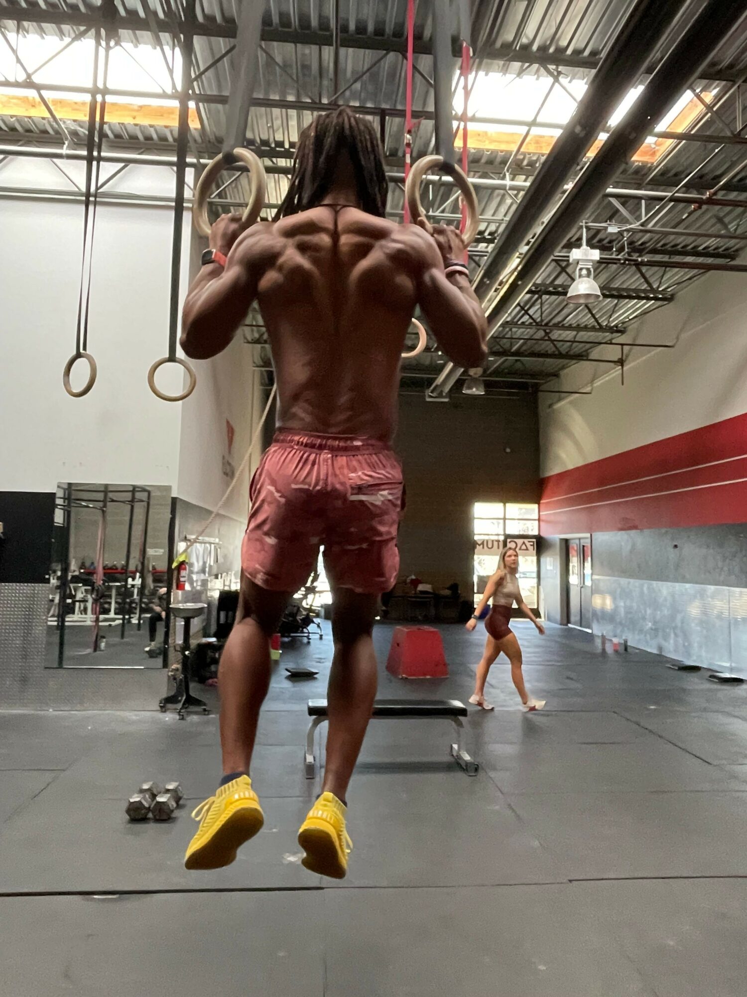 A man with a muscular build and pink shorts is in a gym, stretching with a woman in the background. The gym has metal equipment and a concrete floor.
