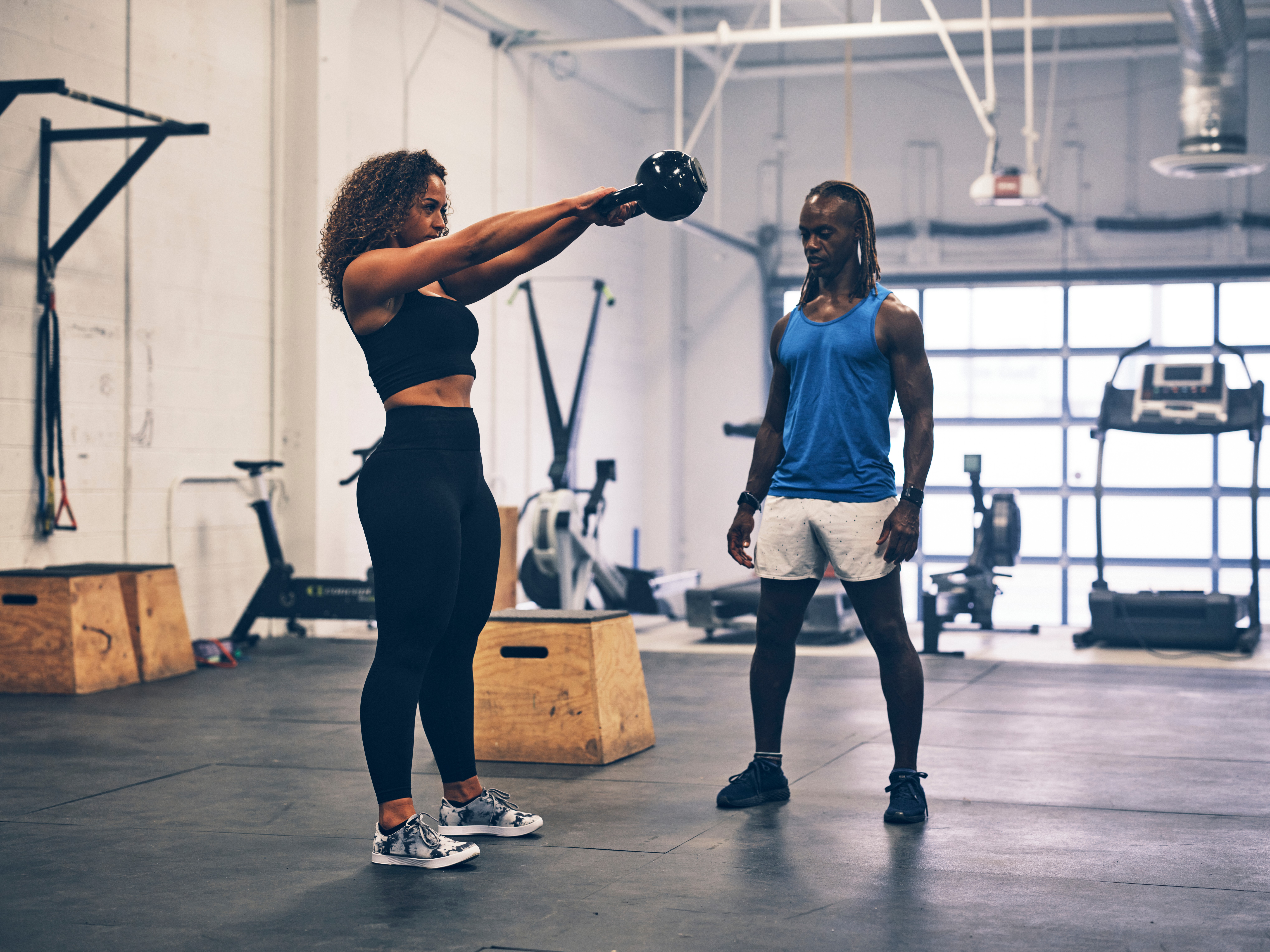 Two women in a gym, one holding a kettlebell with arms extended, the other watching. Gym equipment in background.