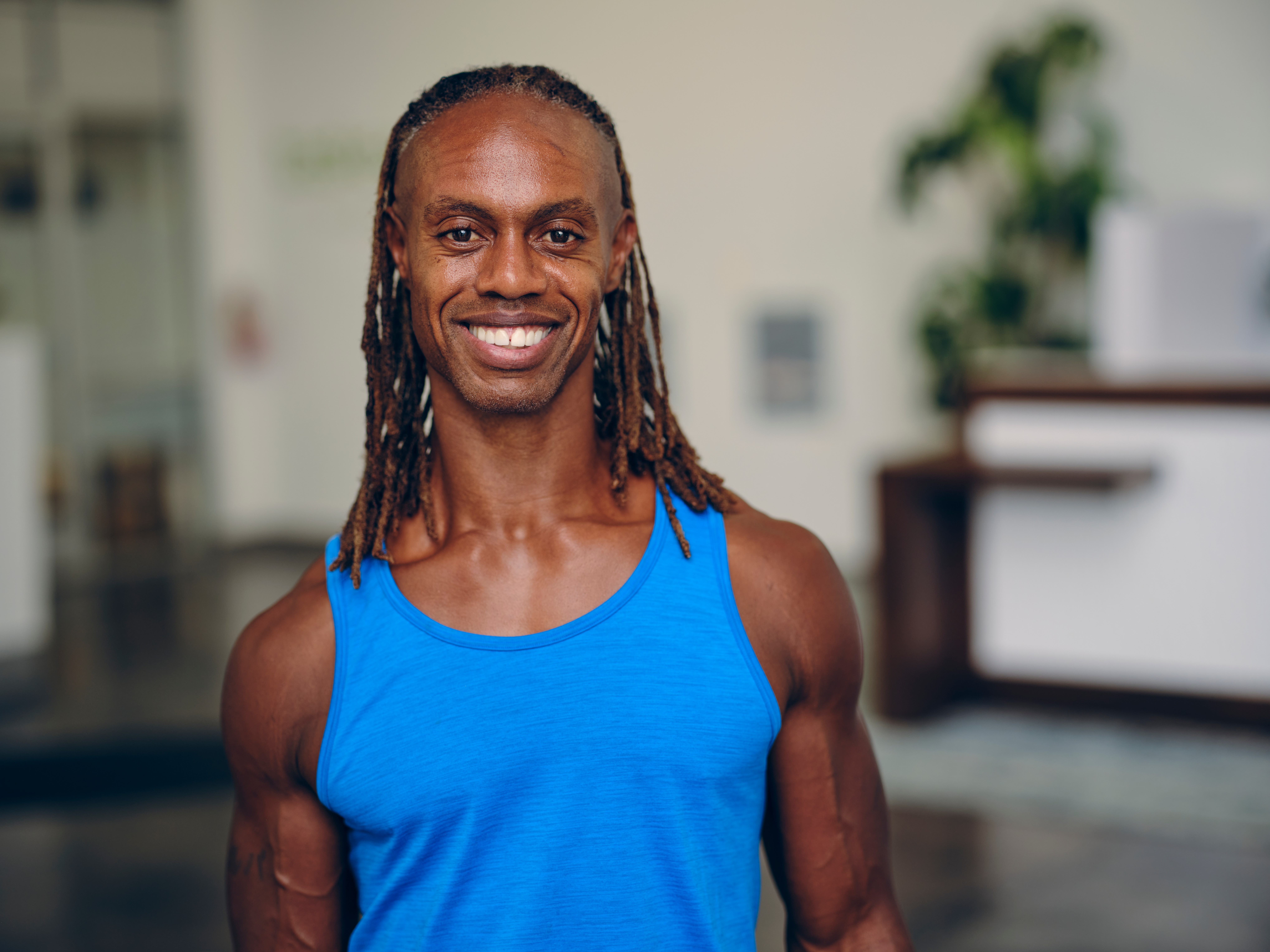 Smiling man with long dreadlocks wearing a blue sleeveless shirt indoors.