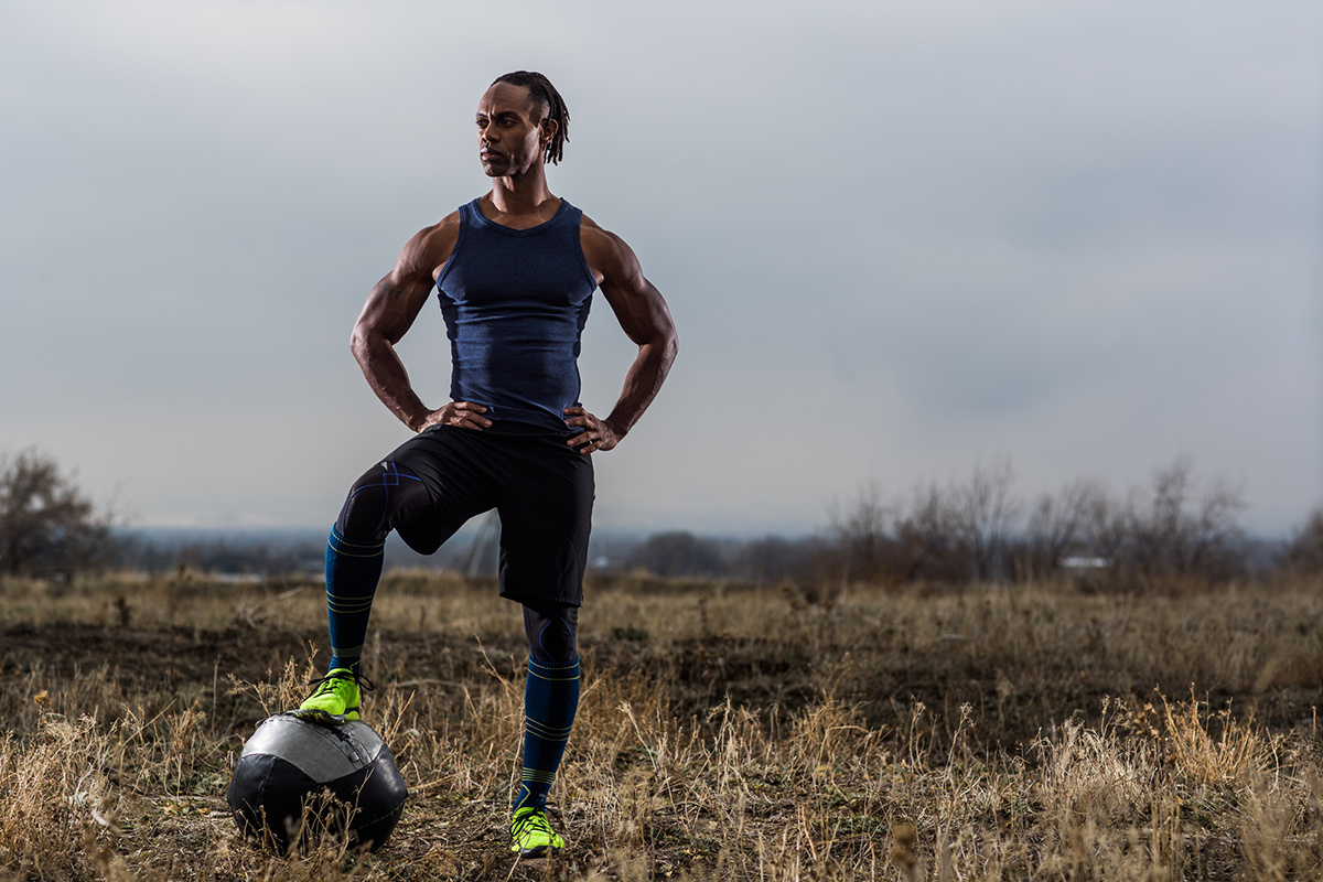 Athletic man standing outdoors with hands on hips, one foot on a black and white soccer ball, overcast sky background.