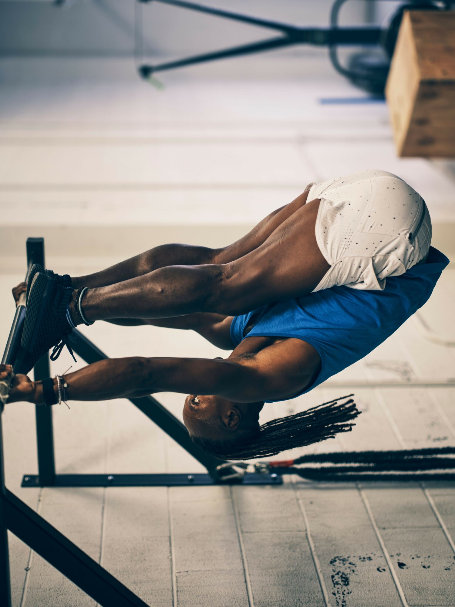 Person hanging upside down from a bar, legs bent, in a gym setting, wearing a blue shirt and white shorts.