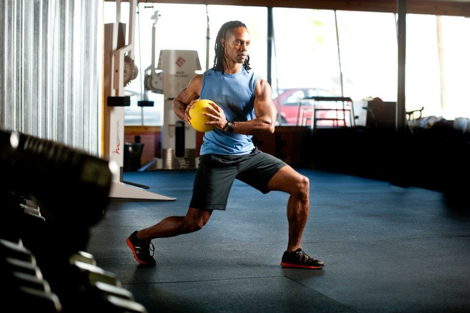 Person exercising with a yellow ball in a gym, lunging forward with one leg, wearing a blue tank top and black shorts.
